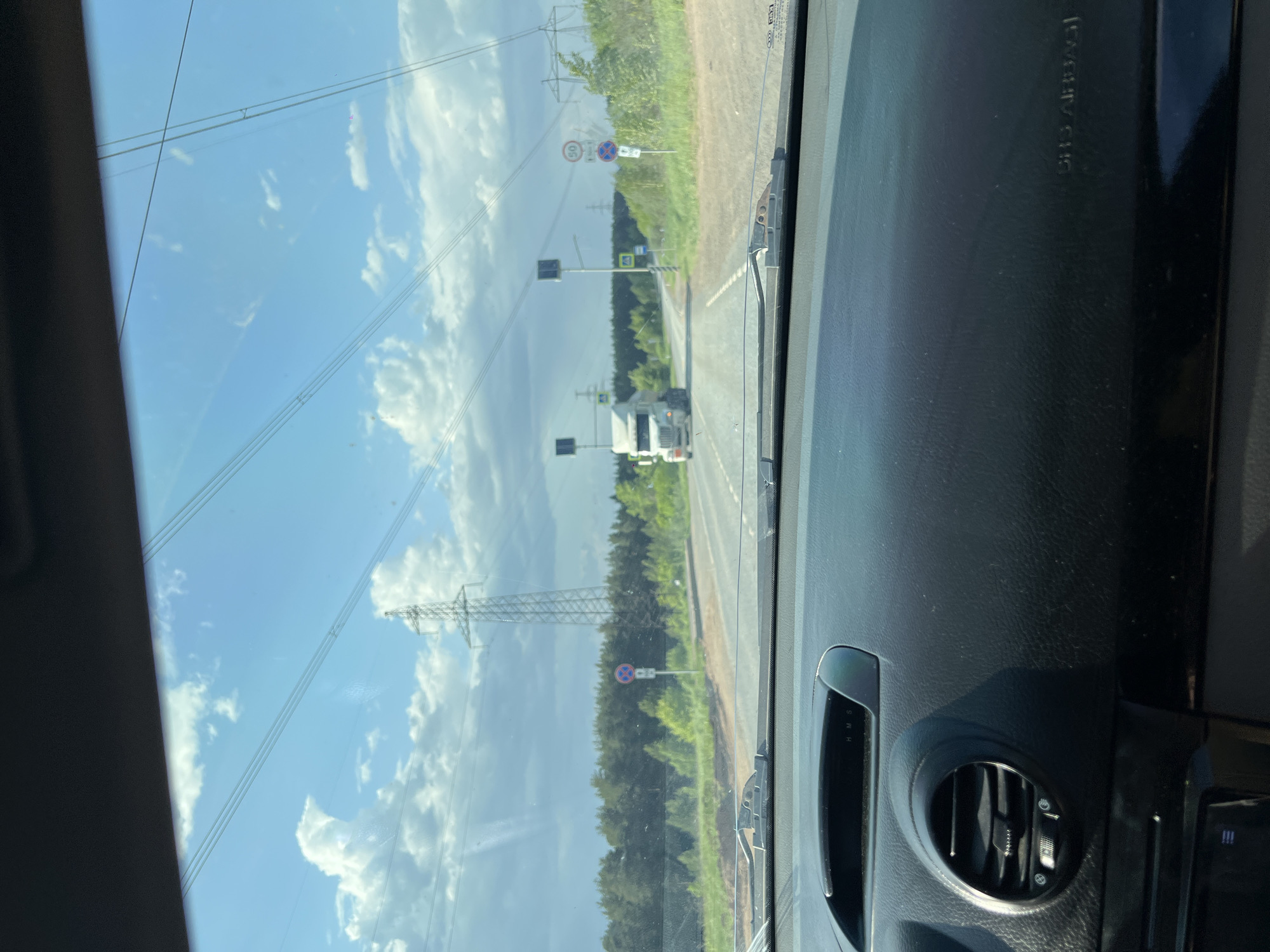 The image, taken from the interior of a car, shows a road with a white truck driving on it. The background includes a green treeline, power lines, and a sky with clouds. The location appears to be near the city of Chaikovsky, Russia, evidenced by the presence of road signs written in Russian, speed limit of 50 km/h and the vehicle’s left-hand drive. The dashboard of the car is visible in the foreground.