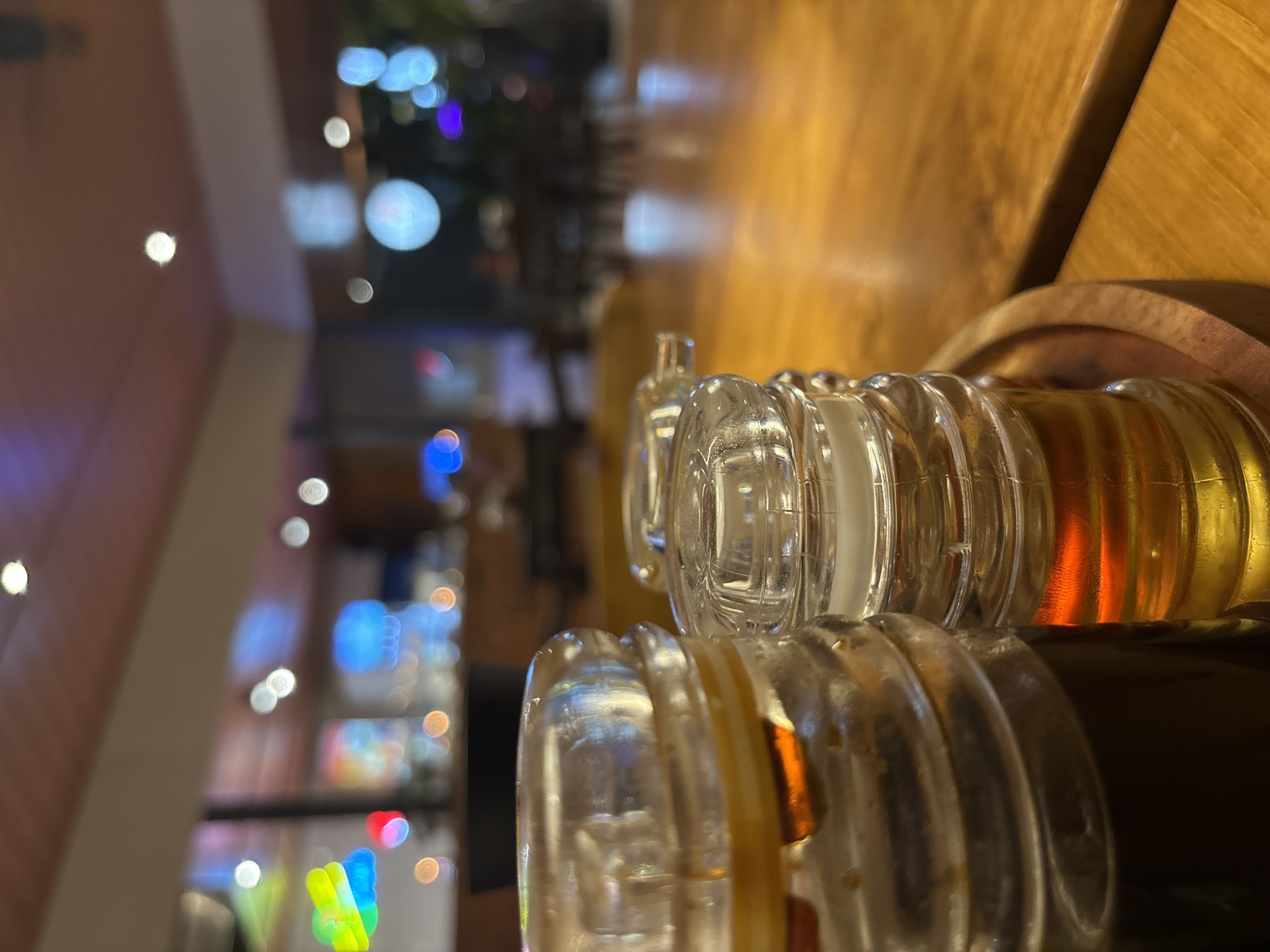 The image captures a close-up view of two glass condiment dispensers on a wooden table. The dispenser on the left contains a dark liquid, likely soy sauce, while the one on the right holds a lighter, amber-colored liquid, possibly cooking oil or a similar dressing. The dispensers have ribbed glass bases and clear plastic lids. The background is blurred but suggests an indoor setting, possibly a restaurant, with soft lighting and out-of-focus colorful lights indicating a lively atmosphere. The wooden table surface shows a warm, polished finish. The scene is forensic in its clarity, focusing on the objects without any human presence or activity. The overall impression is one of a quiet moment at a dining establishment. The implied location, Kuta Selatan, Indonesia, suggests a tropical, potentially tourist-oriented setting, though the image itself does not provide explicit visual cues for this specific locale.