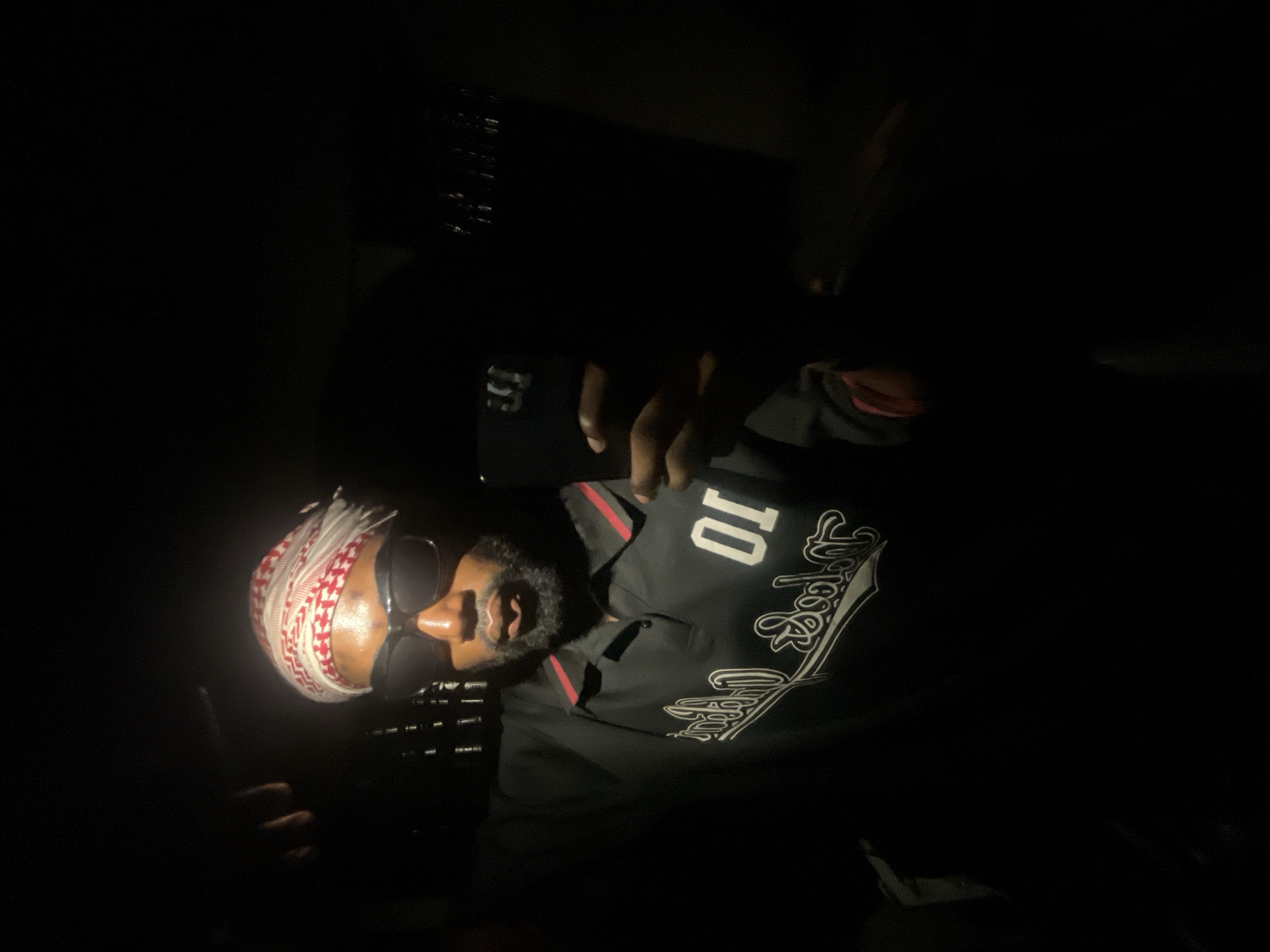 A person with a keffiyeh-like head covering and sunglasses is visible in a dark environment. The individual is wearing a black shirt with a white "Los Angeles" script and the number "10" on it. Part of a hand is raised in the image. The location is unknown and there is no information available about Lokoja, Nigeria. The image is poorly lit.