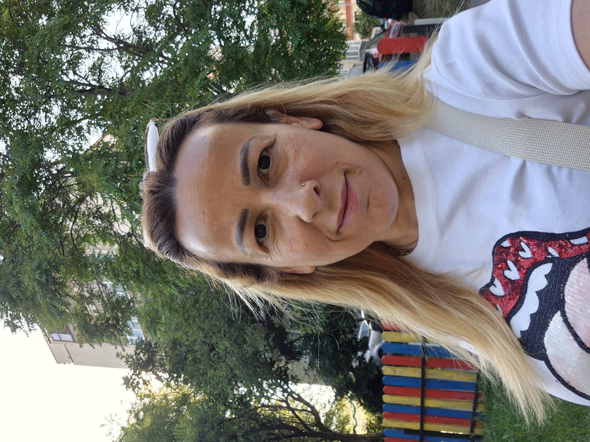 A medium shot photograph shows a woman with blonde hair and light skin, wearing a white t-shirt with a colorful sequined lip design. She is outdoors, with a building and trees in the background. A brightly colored fence is visible to her left. The lighting suggests it is daytime, with sunlight filtering through the trees.