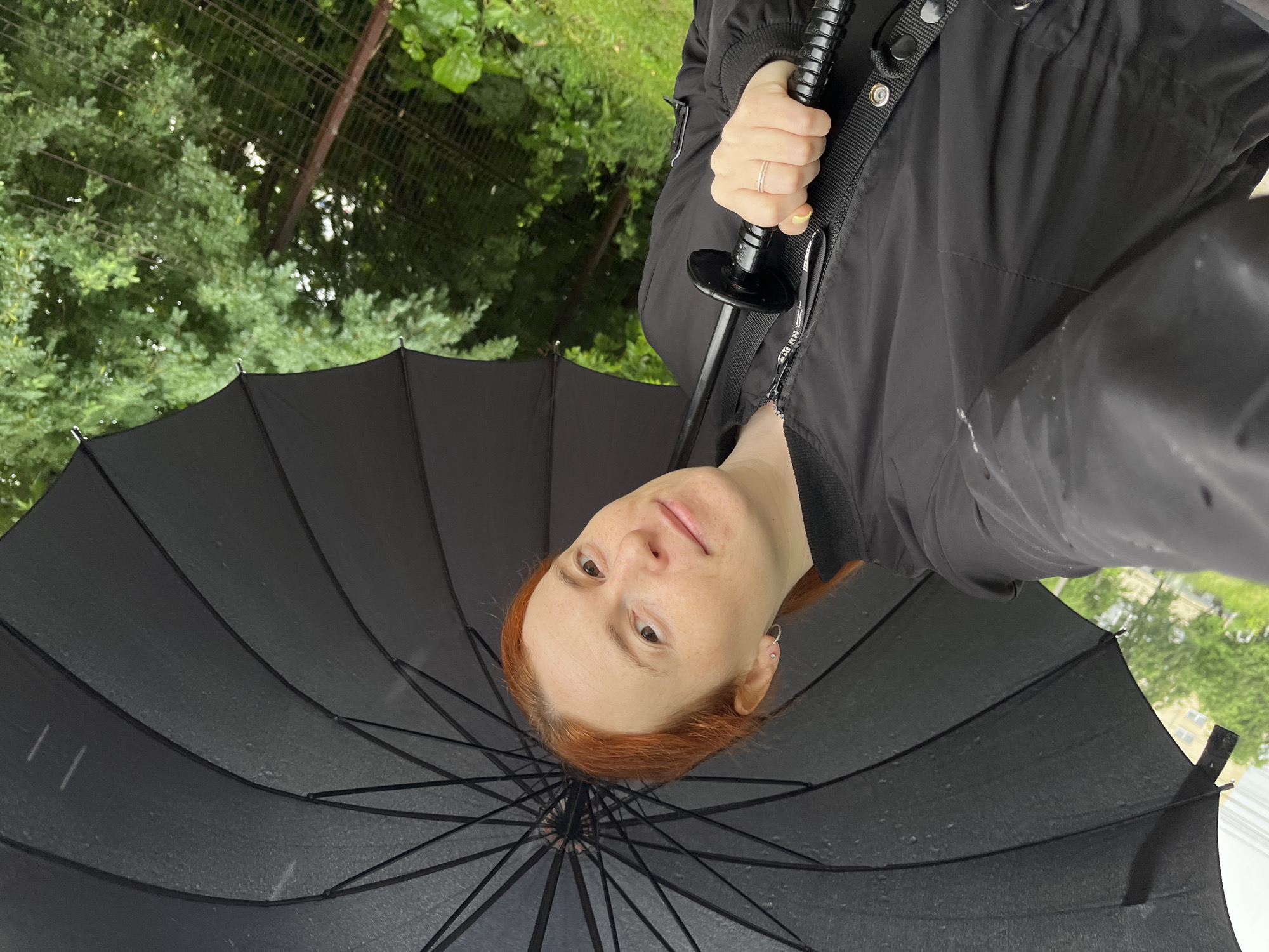 In an outdoor urban environment, possibly a park or residential area in Chaikovsky, Russia, a young woman with red hair and freckles holds a large black umbrella open, shielding herself from the rain. She is wearing a black bomber jacket. The umbrella is partially obscuring her face, but she is looking towards the camera with a slight smile. The background consists of lush green trees and a dark, chain-link fence, suggesting a natural or semi-natural setting within the city. The ground appears wet, and small water droplets are visible on the umbrella's fabric, indicating active precipitation. The lighting is overcast, consistent with rainy weather.