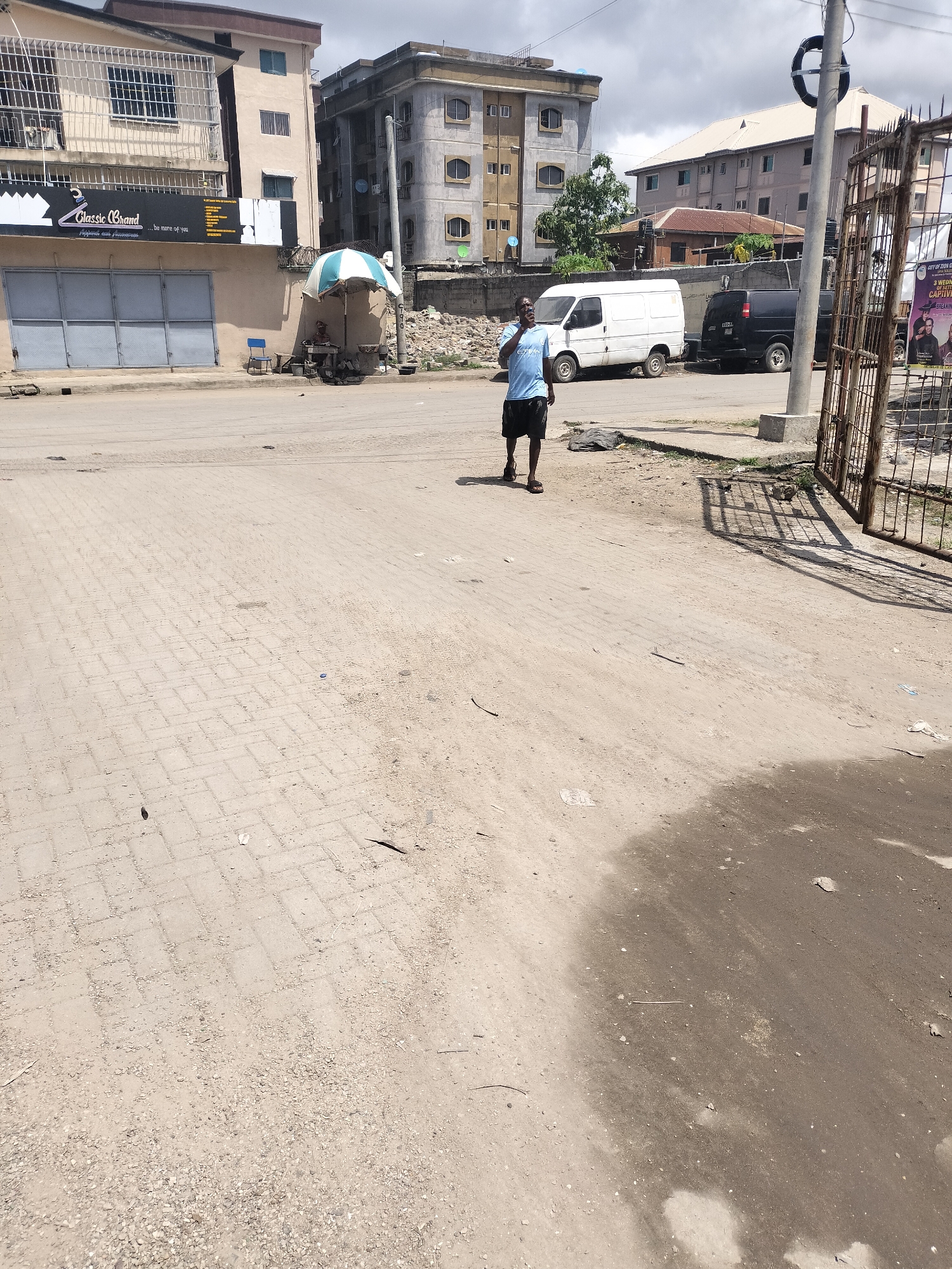 The image depicts a street scene in Alaiyabiagba, Nigeria. The foreground is a dirt and brick road with some debris. A man in a blue shirt and dark shorts is walking in the middle of the road, facing to the left.
In the background, there are several buildings. On the left, there is a storefront with a sign that reads "Classic Brand." A small blue and white striped umbrella stands near a vendor setup. Further back, there are other buildings, one of which is multi-story with grey concrete. There are two vans parked near the street.
To the right of the frame is a metal gate. A poster is attached to the gate with the words "CITY OF ZION G" visible.