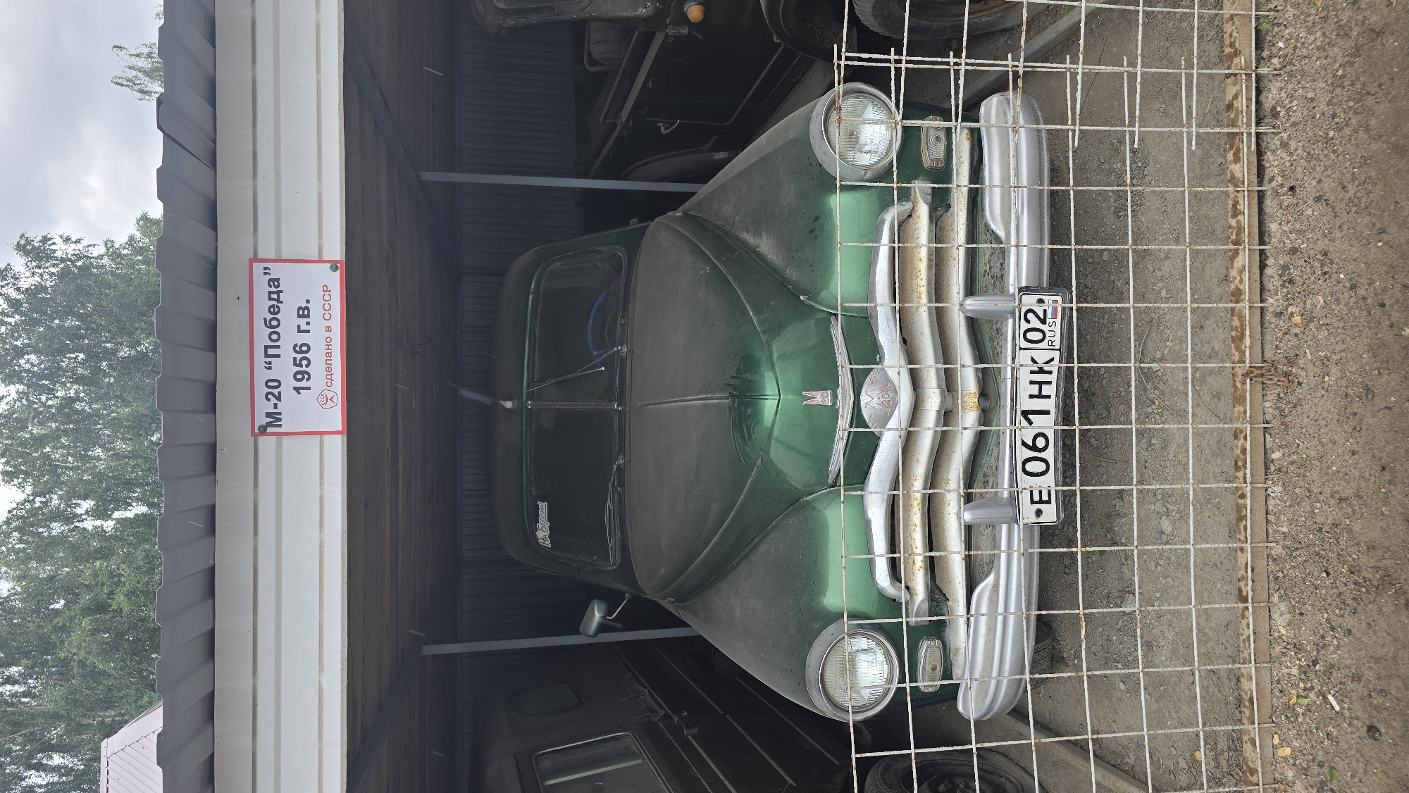 In the city of Bol'shoi Mashlyak, Russia, a vintage green GAZ M-20 Pobeda from 1956 is parked inside a covered, open-air garage. The car is covered in dust, and a white sign with red lettering above it reads "M-20 "Pobeda" 1956 G.V. Made in USSR". The license plate reads "E 061HK 02 RUS". The car is behind a metal fence, and other vehicles are visible on either side. The ground is covered in dirt and gravel.