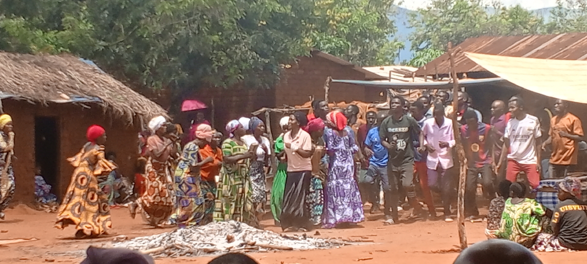 In Ngalula, Democratic Republic of the Congo, a large group of people congregates in an outdoor setting. A group of women are on the left side of the frame wearing bright, patterned dresses and head coverings. Men, some wearing shirts and pants, are to the right. Some people are seated on the ground and are wearing similar clothes. In the background, there are small buildings and trees. The ground is dirt.