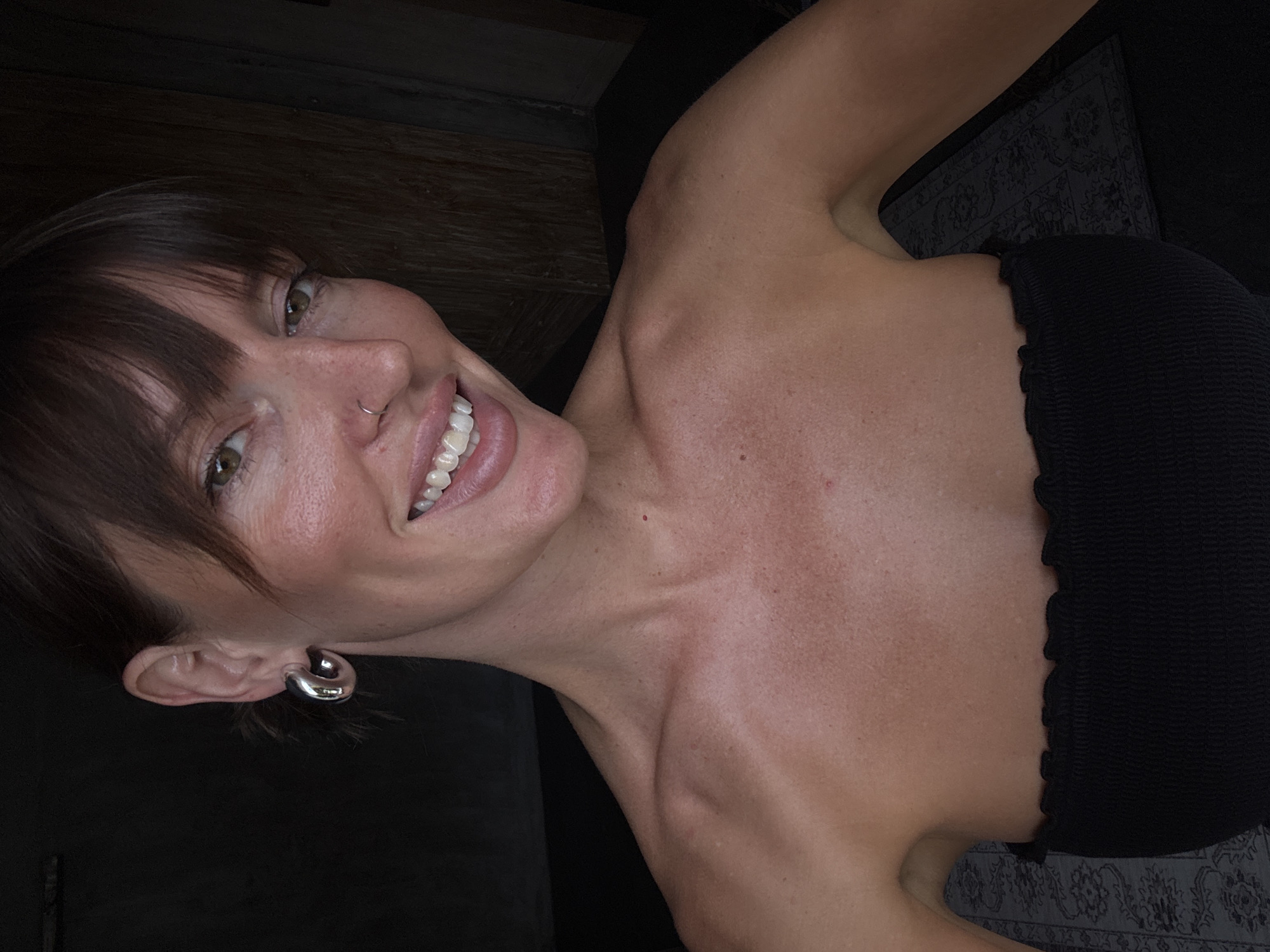 This media file presents a diagonal selfie of a woman, taken indoors. The woman, who has medium brown hair with bangs, is smiling broadly at the camera, revealing her white teeth. Her eyes are open and a shade of green or hazel. She wears a small silver nose ring and a distinctive, thick silver hoop earring in her left ear. Her complexion is fair with visible freckles, and her skin appears tanned. She is dressed in a black, strapless top or dress with a textured, possibly ribbed or ruched, fabric.
The background is dark and indistinct, suggesting an indoor setting. Elements visible include a dark wooden surface or furniture on the left and upper parts of the frame, and a dark patterned rug or textile on the right side, partially obscured by her shoulder. The lighting is soft and even, indicating diffused indoor light rather than direct sunlight. The overall mood conveyed by her expression is joyful and cheerful. No visible text is present in the image. This scene is contextualized as being in North Kuta, Indonesia.
