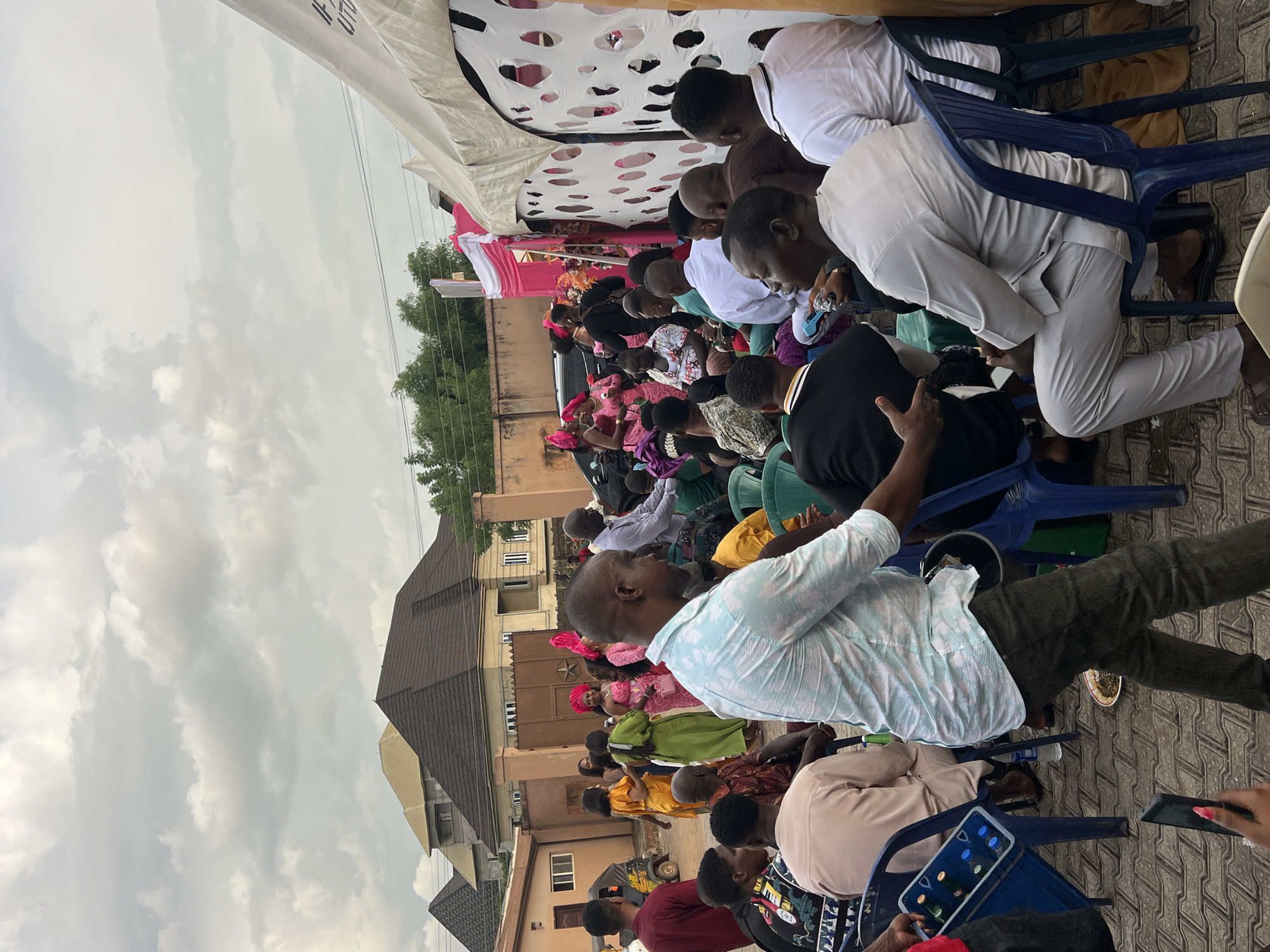 An outdoor gathering of people is taking place in an urban environment. A crowd is seated on blue plastic chairs, facing an area covered by a white tent-like structure with pink accents. The attendees are dressed in a variety of clothing, with some wearing traditional headwear. Buildings with brown roofs are visible in the background, and the sky is overcast.