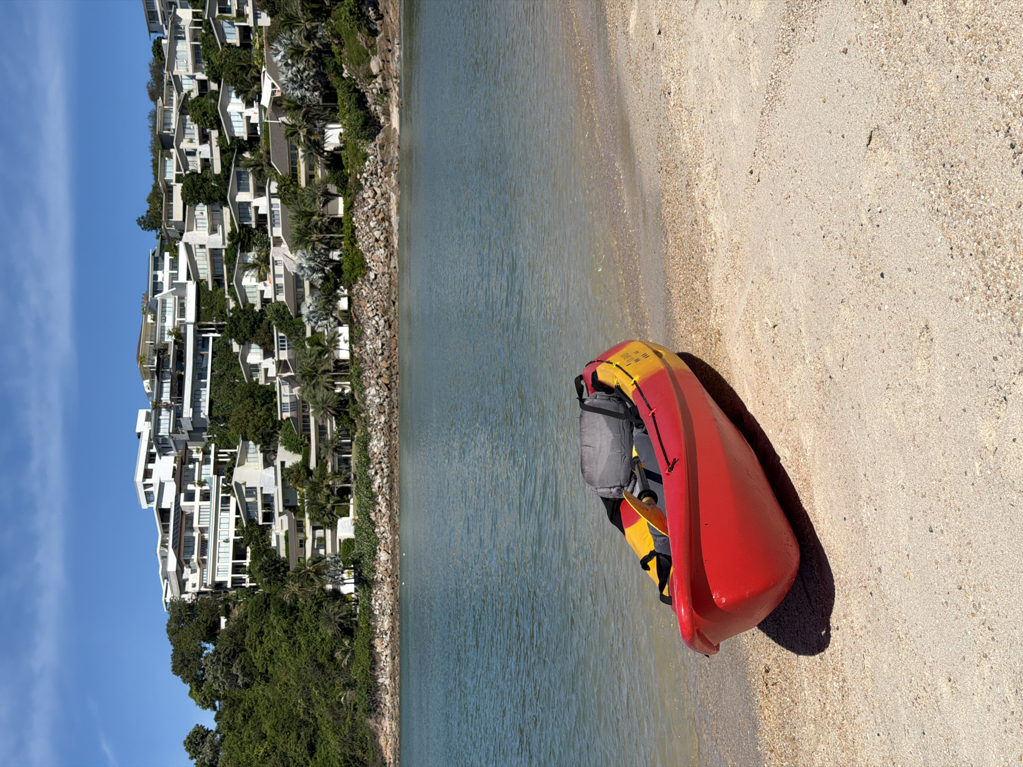 A red and yellow kayak with a gray backpack is resting on a sandy beach. The kayak is positioned diagonally on the sand, with its bow pointing towards the water. The calm, blue water of the ocean stretches out beyond the beach. In the background, a steep, tree-covered hillside is densely packed with modern white buildings, many with balconies and large windows. The architecture suggests a luxury resort or residential complex. The sky is a clear, bright blue with a few wispy clouds. The sun is high, indicating it is likely midday, and the shadows cast by the kayak and the buildings suggest a sunny day. The scene is tranquil and evokes a sense of a tropical vacation. No visible text is present in the image. The location is Baan Plai Laem, Thailand.