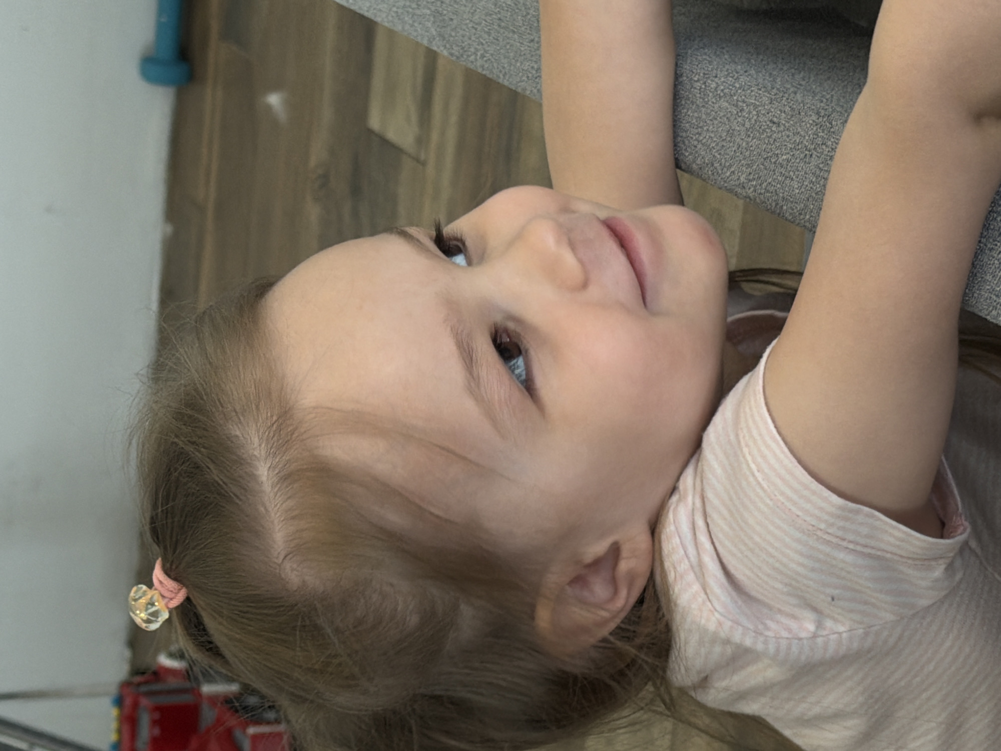 A young child with blonde hair pulled back with a pink scrunchie and a clear heart-shaped bead is looking upwards. The child is wearing a pink and white striped shirt and has their arms raised to hold onto a gray surface. The child's face is turned slightly to the right, and their eyes are visible. The background includes a white wall, brown flooring, and a blue cylindrical object. No indication of the location is visible in the image.