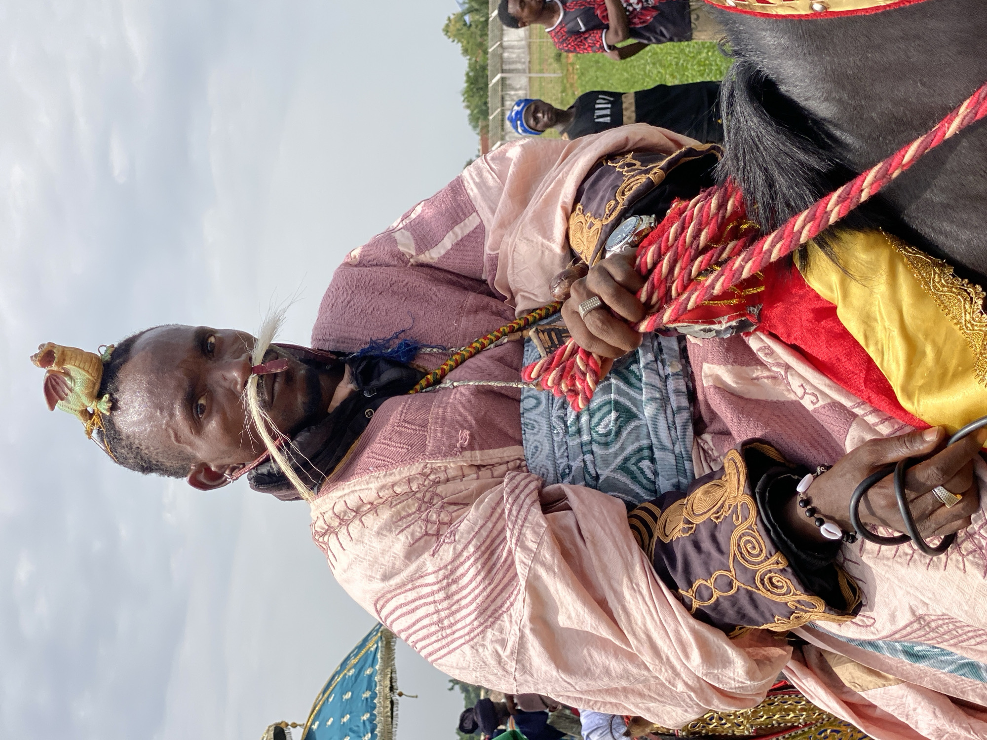 A medium, eye-level shot depicts a man in traditional attire on horseback outdoors. The man has dark skin, short hair, and a thin mustache. He wears a decorative headdress with a small dragon-like figure on the side and a white feather-like object sticking out. His clothing includes a pink and light purple patterned robe, and he wears a ring on his right hand. He holds a red and cream braided rope, presumably reins, in both hands.
A black horse's mane is visible to the man's right. The horse appears to be adorned with colorful fabric, including red and yellow. In the background, several people are visible. There is green grass, a fence, and trees, which suggest a park or open area. The sky is overcast.
The image appears to be taken in the city of Kafanchan A, Nigeria.