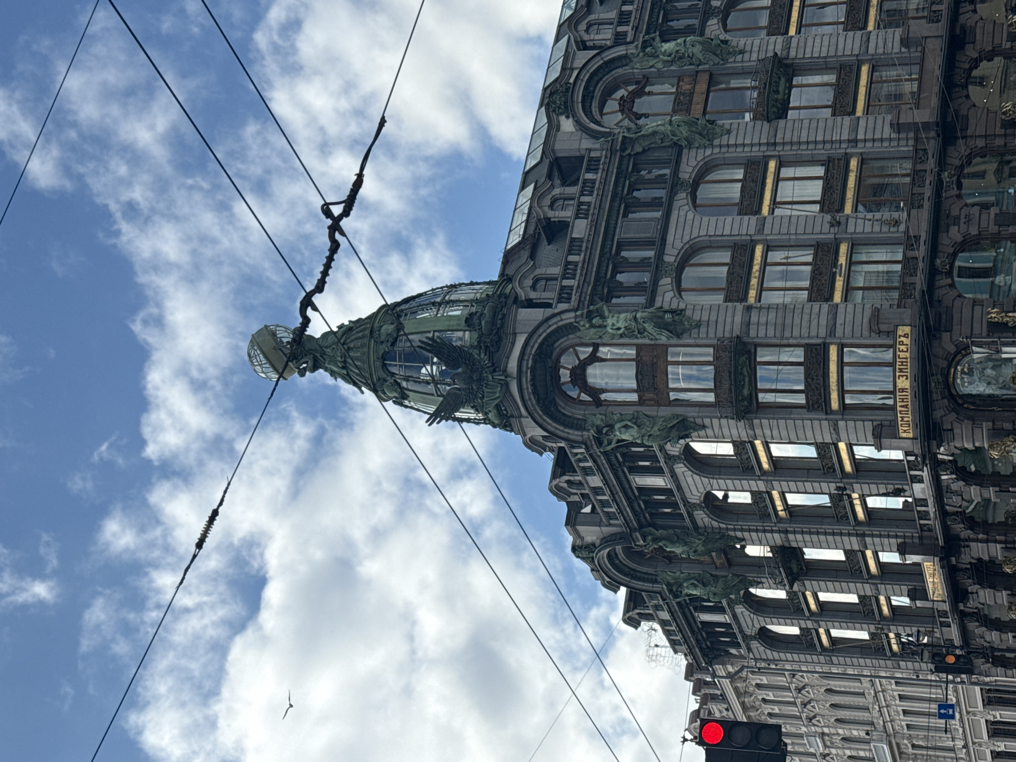 The image captures the architectural detail of the Singer House in Saint Petersburg, Russia, viewed from a low angle looking upwards. The building's facade features ornate carvings, arched windows, and sculptural elements, typical of its Art Nouveau style. Atop the building sits a glass dome supported by bronze figures. Overhead electrical cables intersect the frame against a backdrop of a blue sky dotted with scattered white clouds. A traffic light with a red signal is visible at the bottom edge. A bird flies in the distant sky. Inscribed on the building are the words "КОМПАНИЯ ЗИНГЕРЪ," which translate to "Singer Company."