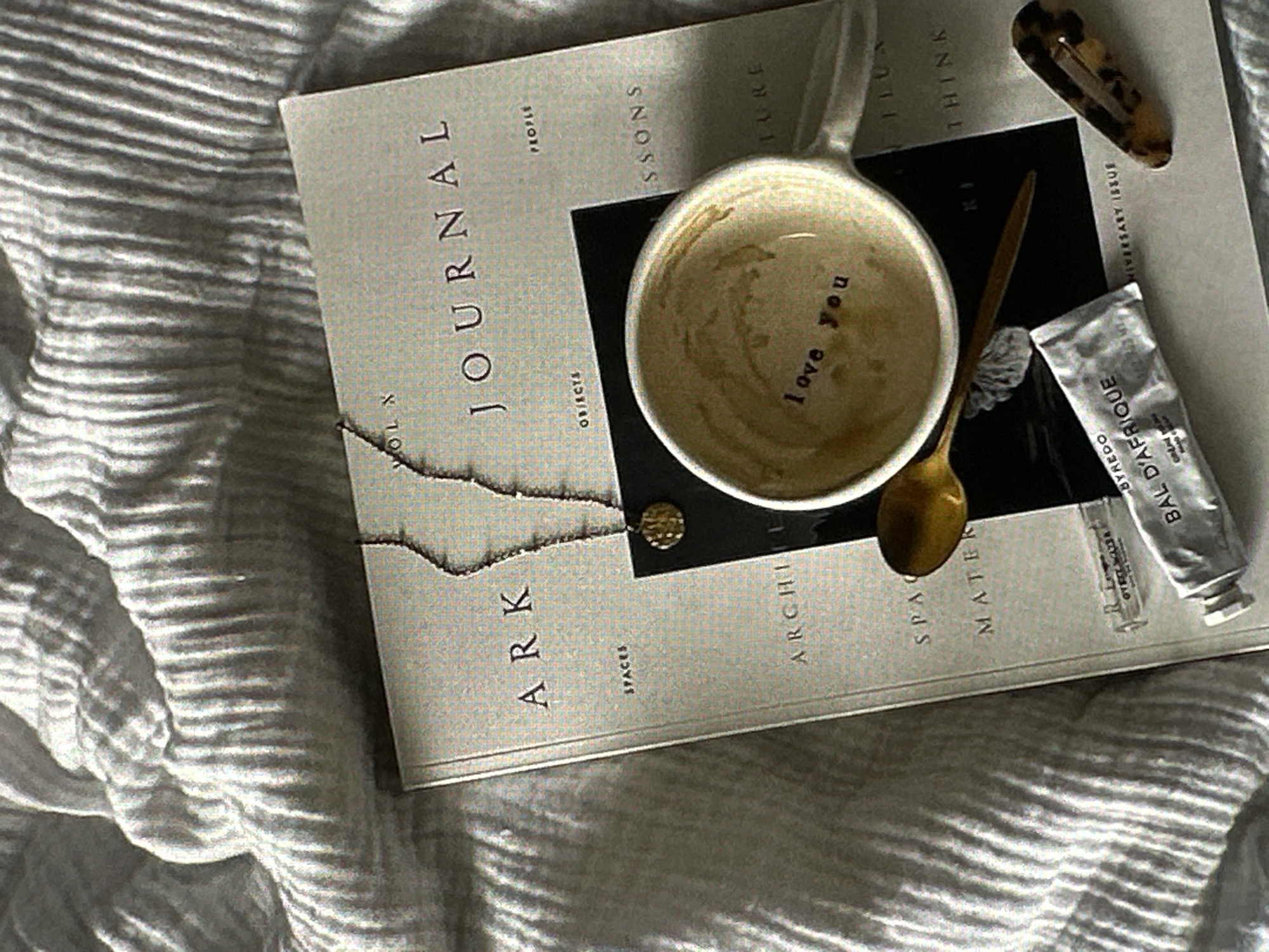 The image shows a book titled "ARK JOURNAL" lying on a striped fabric surface, likely a bedspread or blanket. The book is open, revealing text that includes "SPACES," "ARCHIE," "SPAC," "MATER," "VOL X," "JOURNAL," "OBIECTS," "PEOPLE," "ESSONS," "LURE," and "THINK." A coffee mug with the words "love you" written inside rests on top of the book, along with a small gold spoon. Additionally, there is a Byredo "Bal D'Afrique" hand cream and a hair clip on the book's surface. The environment is soft, neutral, and seems to be indoors, possibly a bedroom. The scene suggests a relaxed and contemplative setting. There is no information about Yerevan, Armenia in the picture.