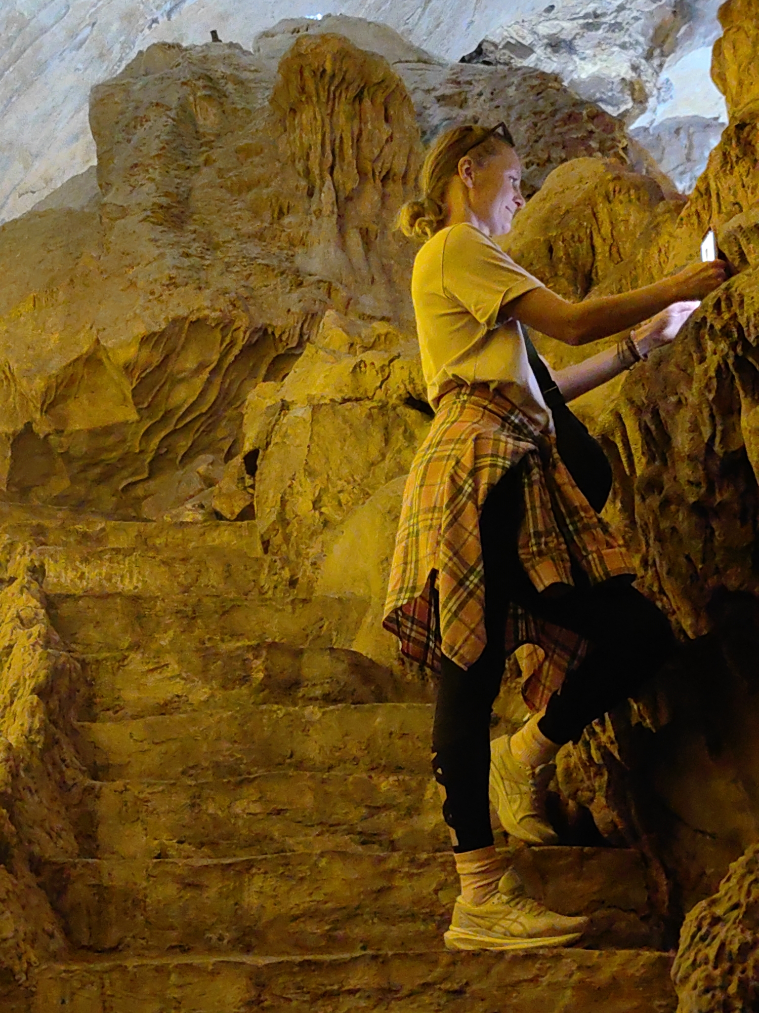 The image shows a woman standing on stone steps inside a cave, possibly a tourist location in Lạng Sơn, Vietnam, based on the cave-like environment. The woman is fair-skinned, has blonde hair, and is wearing a yellow t-shirt, black leggings, and a plaid shirt tied around her waist. She is holding a phone in her right hand and appears to be taking a picture of the cave wall. The cave is illuminated by an unseen light source, highlighting the textured rock formations.