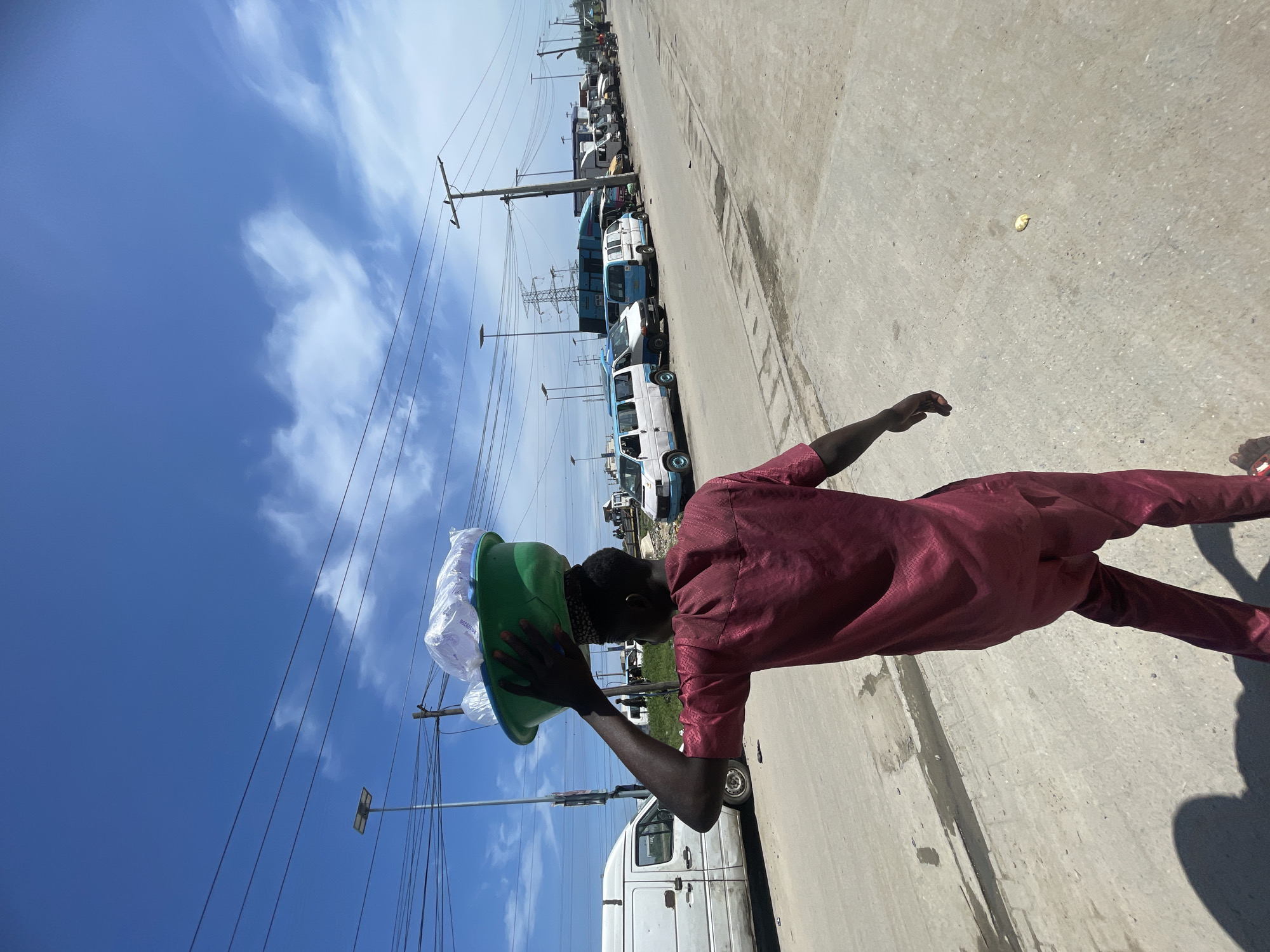 A tilted, eye-level photograph captures a bustling street scene, likely in Lagos, Nigeria, under bright daylight. The primary subject is a person, viewed from behind and slightly to the side, walking along a concrete or asphalt surface. The individual wears a dark red or maroon traditional outfit, comprising a long-sleeved top and trousers, and is balancing a green basin filled with white, translucent packaged goods on their head. One hand is slightly raised, possibly steadying the load.
In the background, a long line of vehicles, mainly white and blue vans and buses, are parked or moving slowly along the roadside, suggesting a transport hub or busy thoroughfare. These vehicles appear somewhat aged. A dense network of utility poles and power lines stretches across the frame, dominating the upper middle ground.
The sky is a clear, vibrant blue with scattered white clouds, indicating sunny weather. Strong shadows are visible on the ground, reinforcing the bright daylight conditions. Faint, partially illegible text and numbers, such as "91-1025B", can be discerned on some of the packaged items in the basin. The image captures a common sight of street vending and urban activity in a vibrant setting.