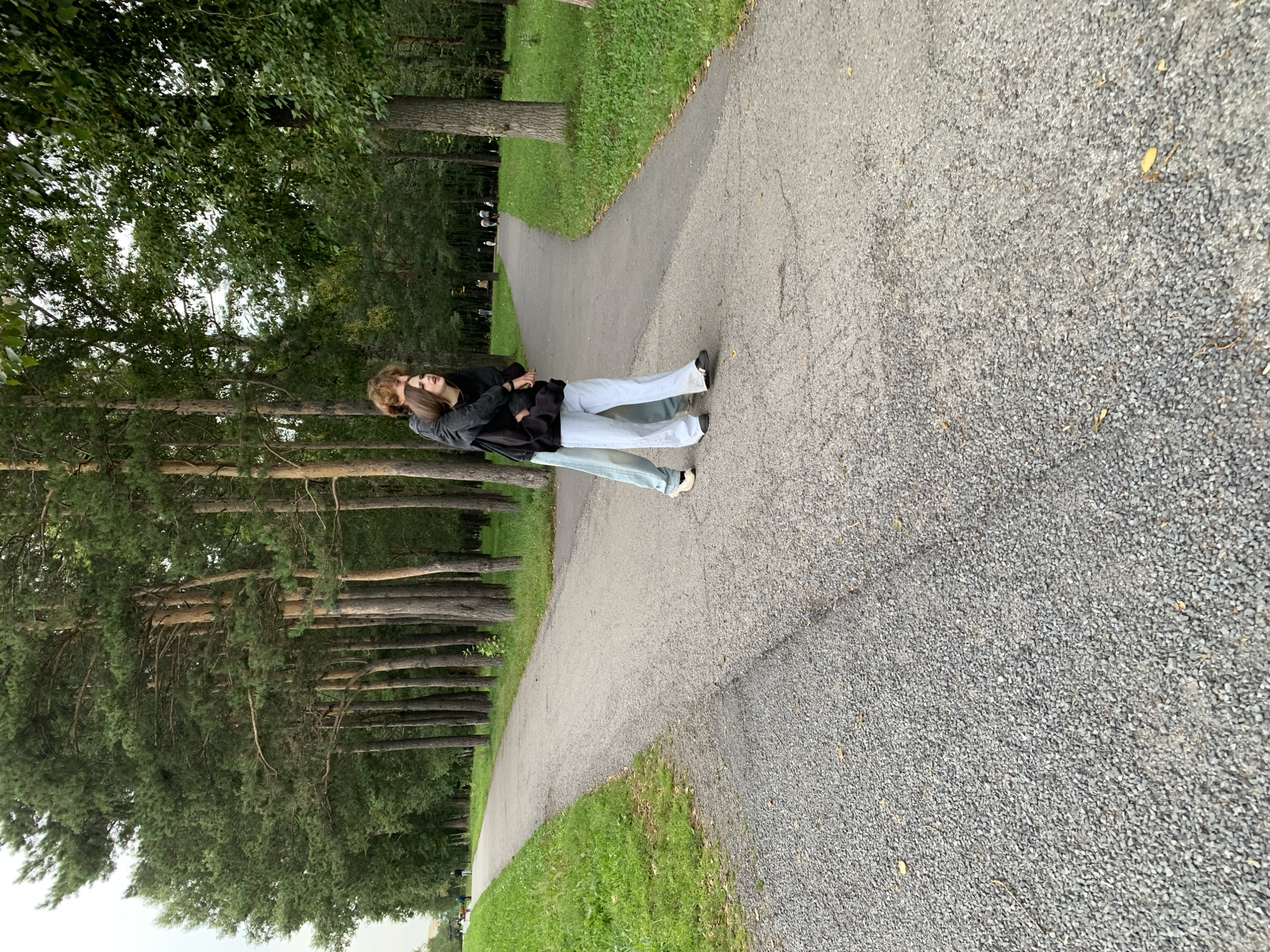 Two individuals are positioned in the center of a paved pathway in a park. The person in the foreground, from the waist down, is wearing light blue pants and black shoes. They are facing towards the right of the frame. Behind them, a second individual is embracing them from behind, with their arms wrapped around the other person's torso. This second individual is wearing a dark-colored jacket over a dark shirt and also light-colored pants. Their head is turned to the right, and their mouth is open. The pathway splits ahead of them, with one section curving to the left and another to the right. The pathway is lined with tall pine trees on the left and a mix of deciduous and coniferous trees on the right. The ground beside the pathway is covered in grass and gravel. In the background, several other individuals are visible on the pathways, and the trees extend into the distance. The sky is overcast. The overall scene depicts a moment of intimacy and connection between two people in a natural, outdoor setting, likely within a city park in Prokopyevsk, Russia, suggested by the presence of well-maintained pathways and diverse tree species common to the region.