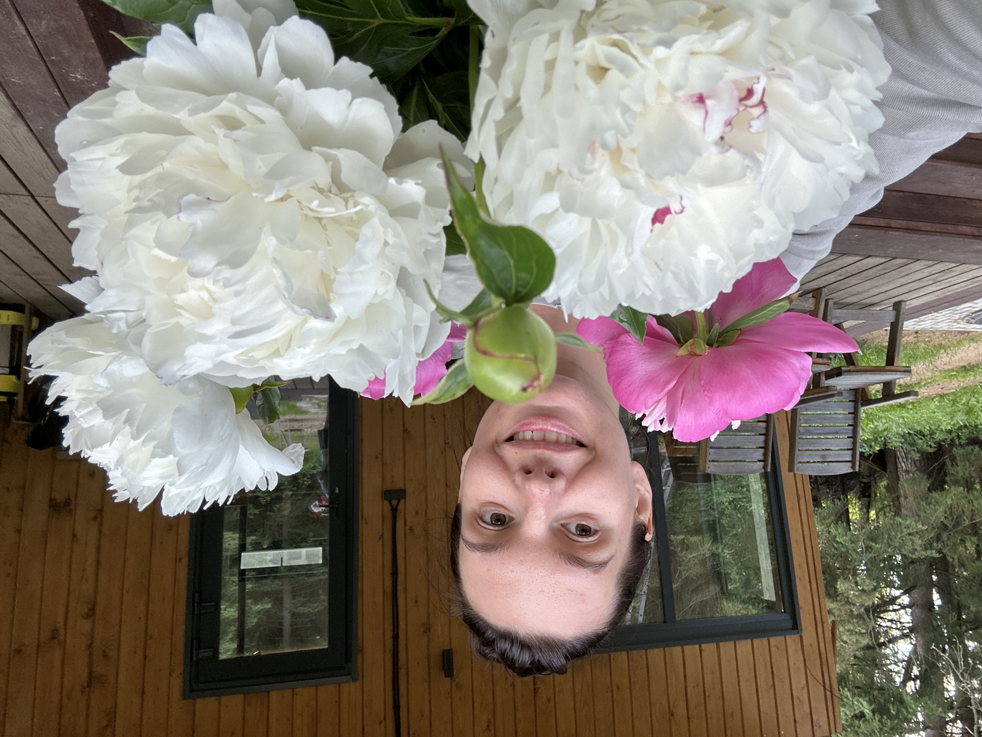 A medium shot photo shows a woman holding a bouquet of peonies. She is smiling directly at the camera. She is wearing a grey t-shirt. The background shows a wooden house with a large window and a door. To the left of the house are two wooden chairs and trees. The woman is in the foreground of the image, with the peonies partially obscuring her face. The location is Прикамский, Russia.