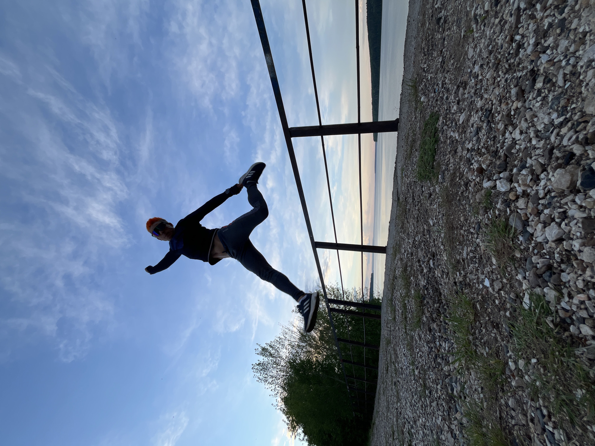 The image depicts a person performing a yoga pose near a railing. The person is wearing athletic clothing, including a jacket and leggings, an orange hat, and sunglasses. They are positioned sideways, appearing to balance on the railing with one hand while extending the other arm and leg in a yoga posture. The background shows a body of water, likely a river or lake, with land visible in the distance. Above, there is a partly cloudy sky. The scene also includes a gravelly or rocky ground and some greenery. The location is identified as being in the city of Chaikovsky, Russia.