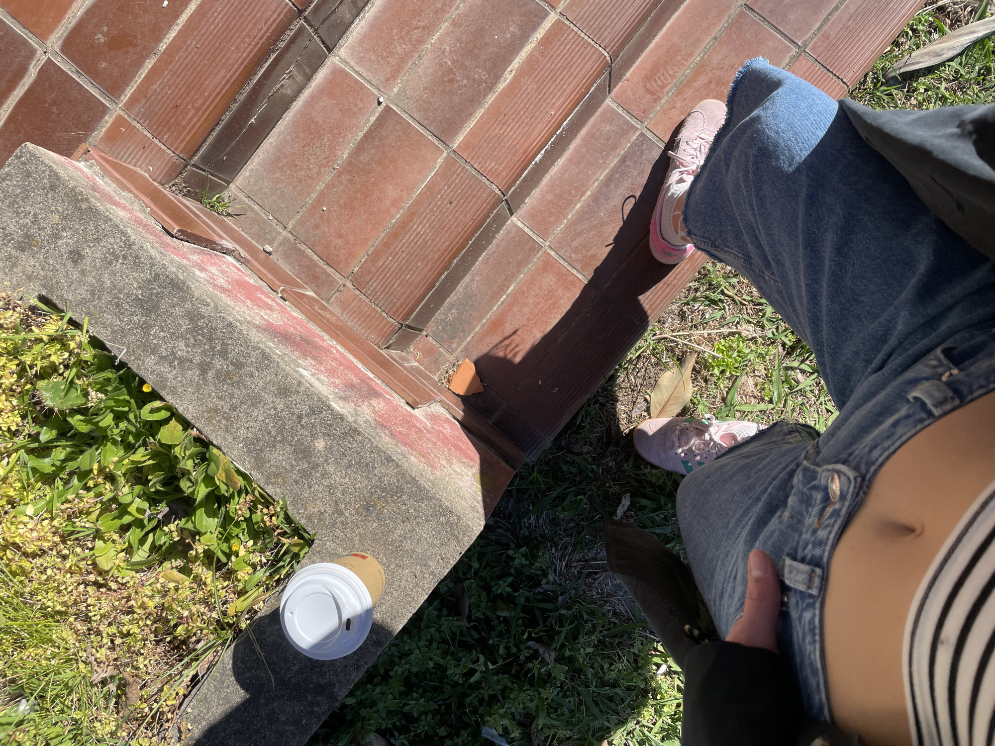 The image depicts a first-person perspective of a person standing on a brick walkway adjacent to a concrete ledge with vegetation. The person is wearing light blue jeans and pink sneakers. A coffee cup sits on the ledge near the vegetation. The location is unknown and not visibly indicated in the image. The image does not show a discernible cityscape or landmarks pointing to the city of Budva, Montenegro.