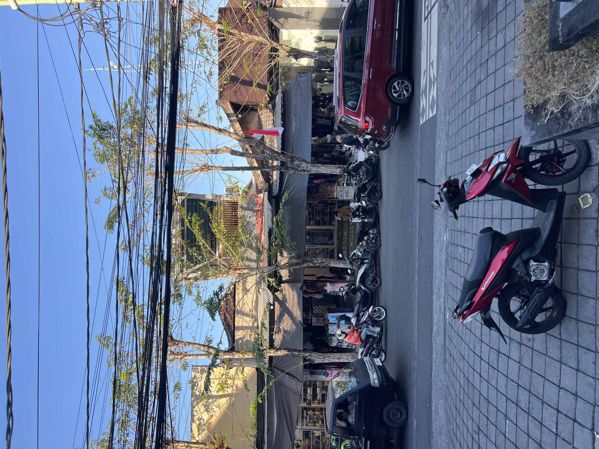 A street scene in Kuta, Indonesia shows a paved road with a red scooter parked on the side. In the background, several shops are open, displaying clothing and other goods. People are seen walking and riding motorcycles, and a red van and a black truck are parked on the street. Trees with sparse foliage line the street, and a multitude of electrical wires are strung overhead. The sky is clear and blue. The overall impression is of a busy, commercial area on a bright day.