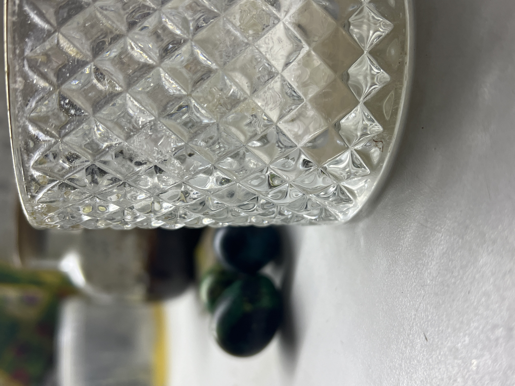 The image shows a close-up of a clear glass with a textured diamond pattern. Below the glass, there are blurry, dark-colored objects with a hint of green. The background consists of a white surface, and some out-of-focus items in the distance.
This image does not provide information about location, people, actions, interactions or environment for it to be in Noviy, Russia. It is a macro photograph of objects on a surface.