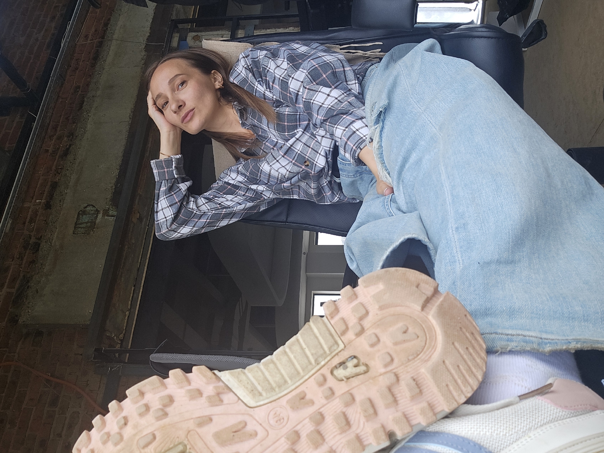 A woman is lying on a black leather-like sofa. She is wearing a grey and white plaid shirt and light blue jeans. Her right hand supports her head. The camera is oriented to capture the bottom of a pink and white shoe in the foreground. The background shows parts of a brick wall, a metal frame structure, and a window. The location is implied to be in the city of Chaikovsky, Russia.