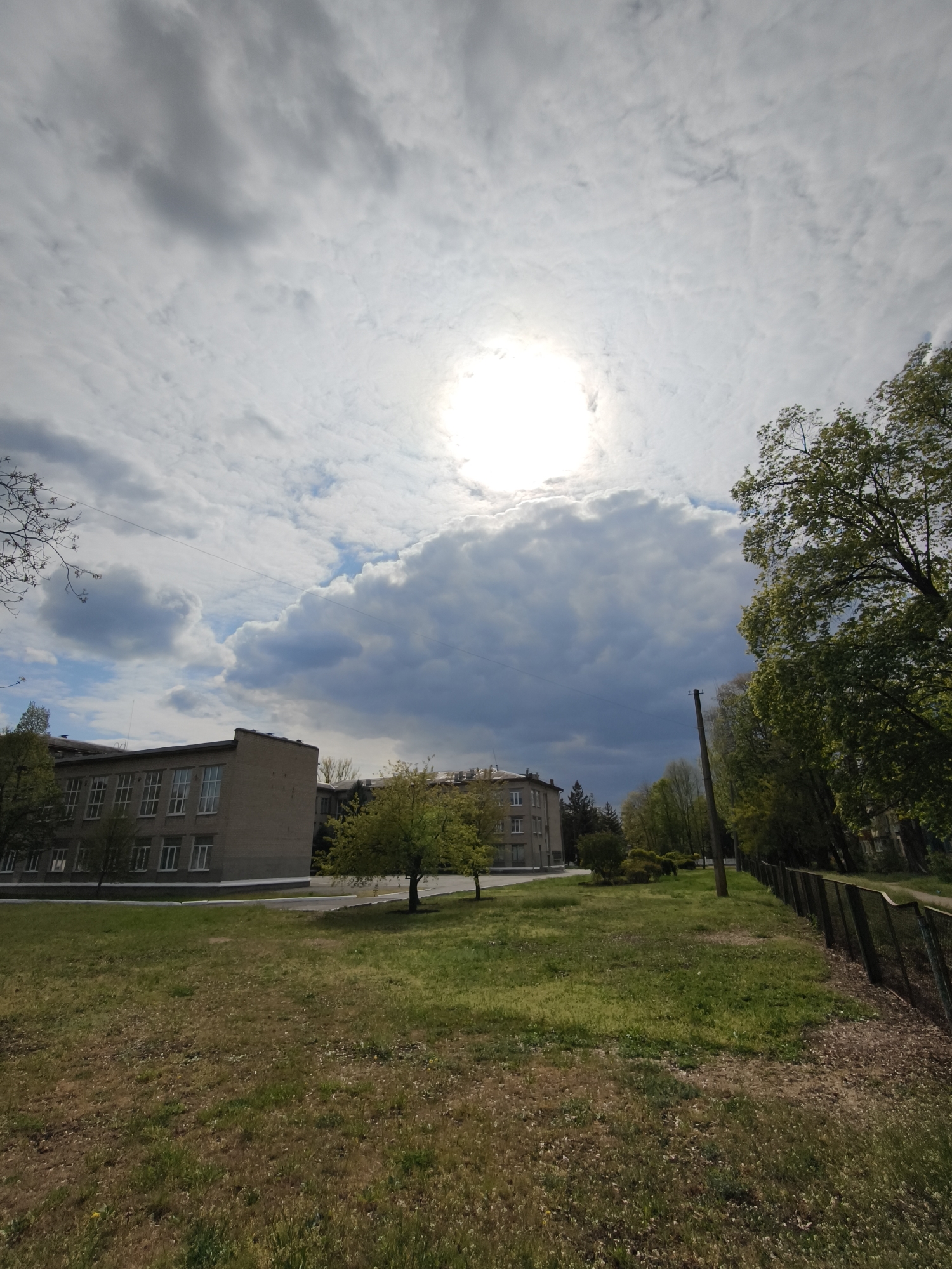 This vertical image captures a tranquil outdoor scene in what appears to be a semi-urban or institutional area, potentially in Kryvyi Rih, Ukraine. The sky is dominated by a bright, intense sun partially obscured by white and light grey clouds, suggesting a mix of sunny and overcast conditions. To the right, darker, heavier clouds are visible, adding to the dynamic weather pattern.
In the mid-ground on the left, there is a multi-story brick building featuring numerous rectangular windows, indicative of a functional or public architectural style. Another similar building is partially visible further back. A paved surface or road runs in front of these structures.
The foreground is comprised of a large, uneven grassy field displaying varying shades of green and some brown patches, suggesting early summer or slightly dry conditions. A small tree with vibrant green foliage stands prominently in the middle of this grassy area. Along the right side of the image, a dense line of mature trees with full green leaves creates a natural border, running parallel to a dark mesh fence. A dark utility pole with an overhead wire is also visible on the right. No people or specific activities are depicted in the scene, which conveys a calm, daytime atmosphere. There is no visible text.