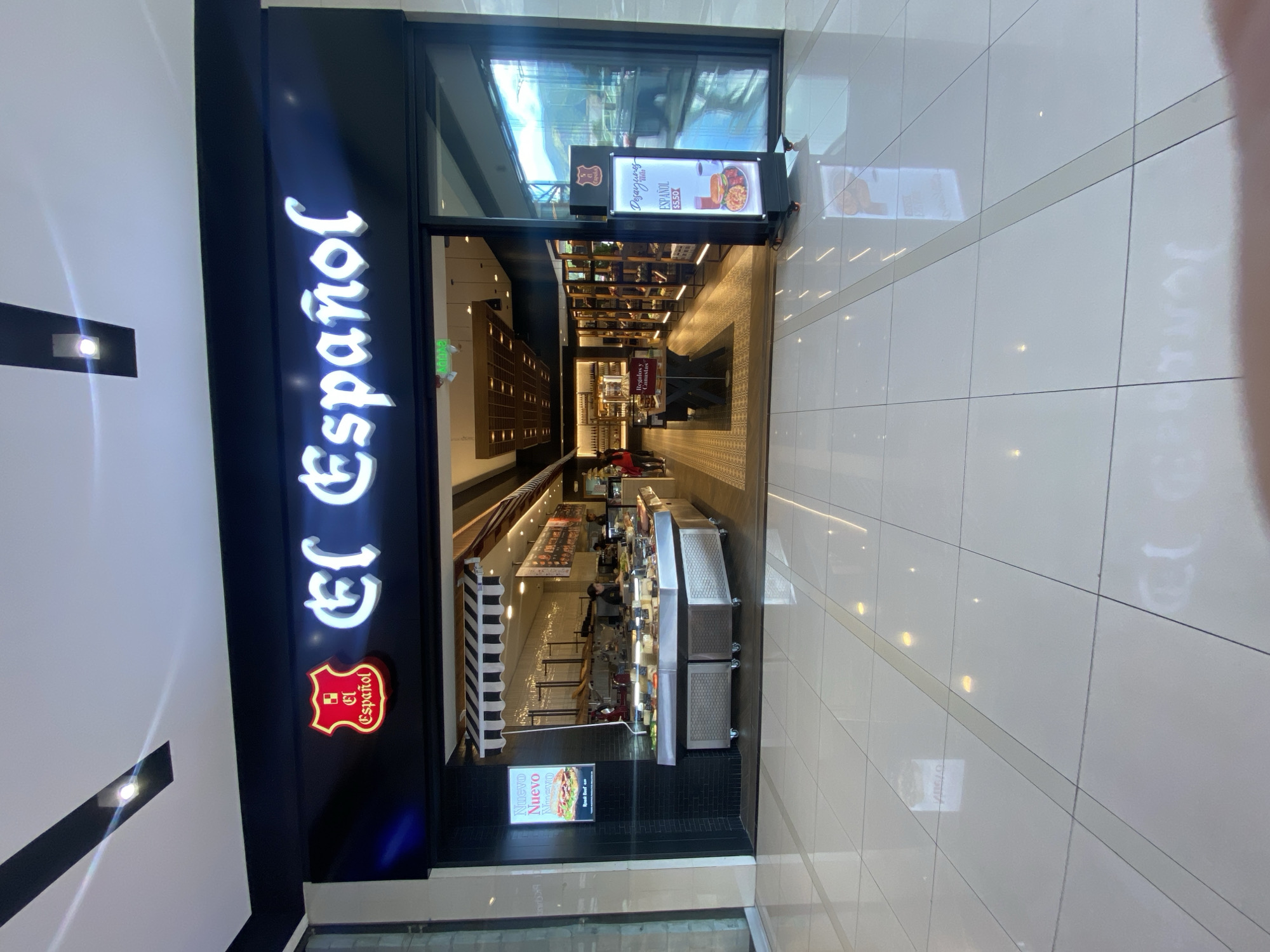 The image shows the entrance of "El Español" a restaurant in Quito, Ecuador. The restaurant's sign is prominently displayed vertically, illuminated in bright white against a dark background. Another sign above the entrance advertises breakfast (desayunos) for $5.50, and a smaller sign displays "Nuevo Nuevo Nuevo Roast Beef". The view extends down a long, narrow space, suggesting a corridor or mall environment, with the restaurant located to the left. The floors are tiled in white. The interior of the restaurant features display cases and a serving area. There are people inside the restaurant. The image is taken from a slightly elevated perspective looking down towards the establishment.