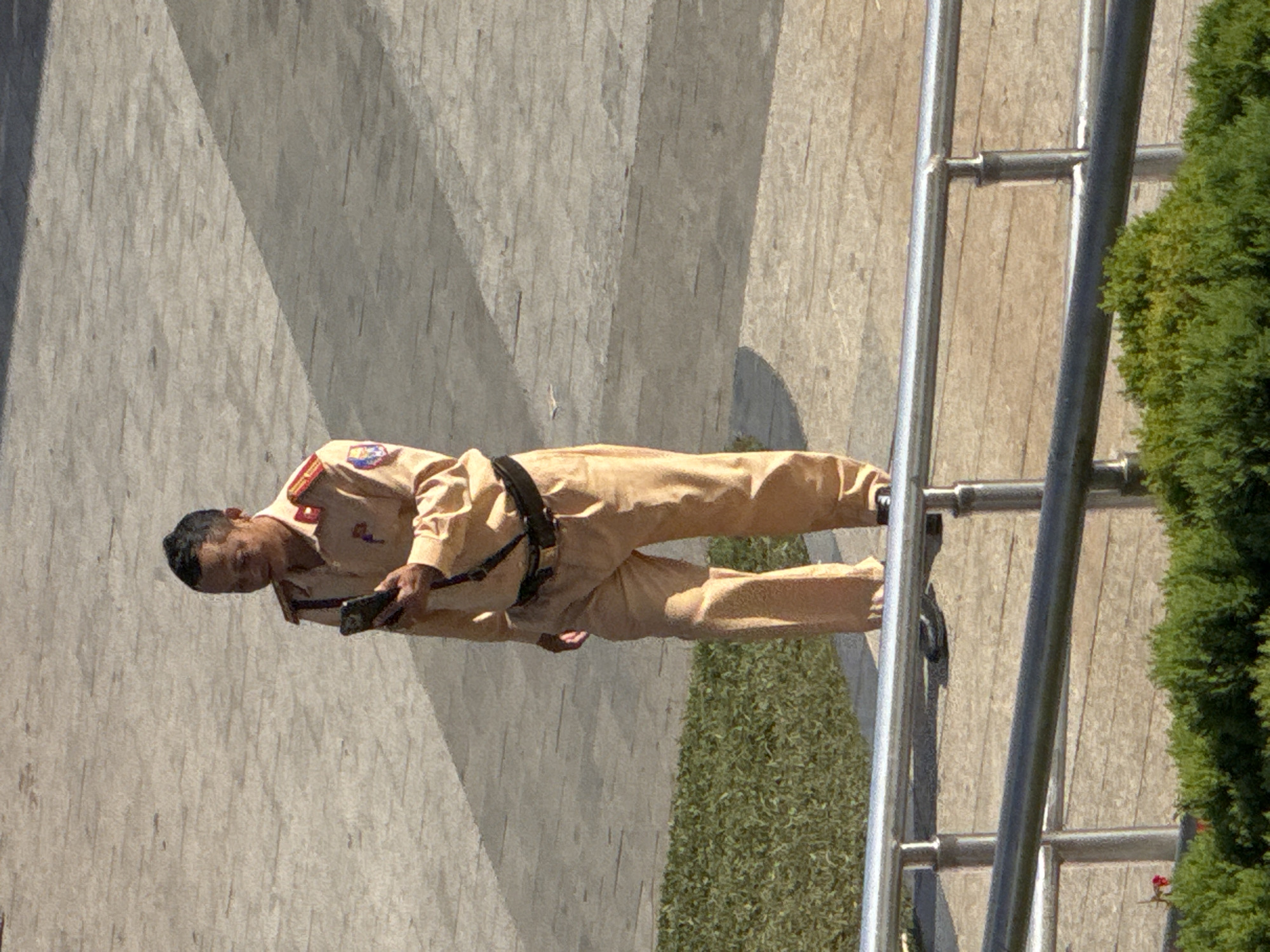 A lone man in a light tan uniform with red accents on the collar and epaulets is standing outdoors on a paved surface. He is looking down at a black mobile phone in his right hand. The man has short dark hair and is wearing a belt. The paved area appears to be a walkway or plaza, with a strip of green grass to the left of the man and a metal railing in the foreground. A dark shadow is cast across the paving stones from the right side of the frame. The background is out of focus. The man's uniform suggests he may be a traffic officer or security personnel, possibly in Vietnam, given the context of Da Lat.