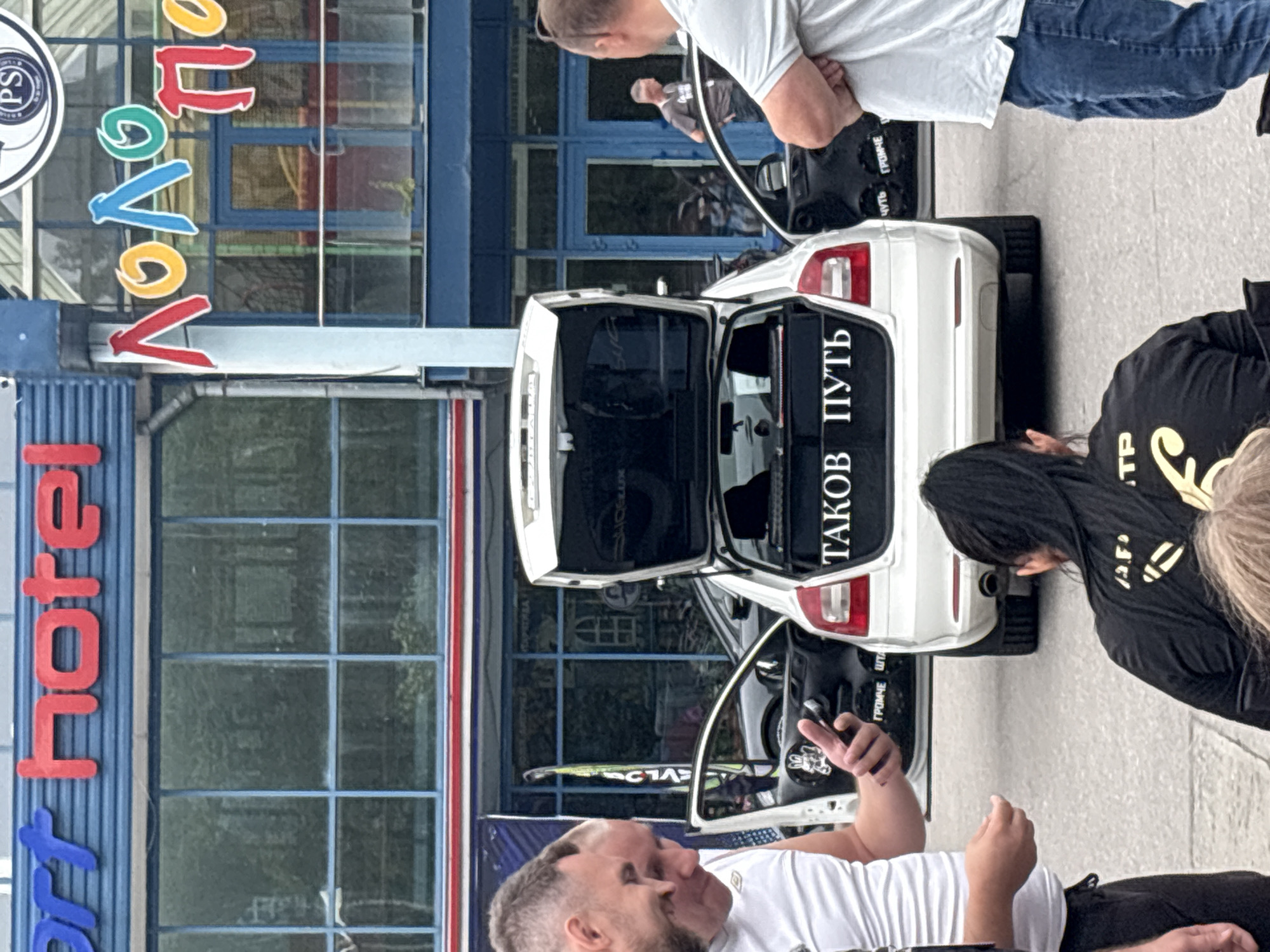 In Zanevka, Russia, a white Skoda Octavia with its trunk and driver's side door open is parked on an asphalt surface. The trunk is fitted with a large audio system, and the rear window has the Russian text "ТАКОВ ПУТЬ" (This is the way) displayed in white lettering against a black background. Several people are gathered around the car. Two men in the foreground are looking at a smartphone held by one of them. A woman with long dark hair is seated in front of the car, facing away from the camera. Another man, wearing a grey t-shirt and jeans, stands near the open driver's side door, looking into the car. In the background, a building with blue and red signage is visible. The signage on the building includes "ort hotel" in blue and red, and colorful Cyrillic letters forming "ЛОЛО ПО". The overall scene appears to be a casual gathering, possibly related to a car audio event or a car show.