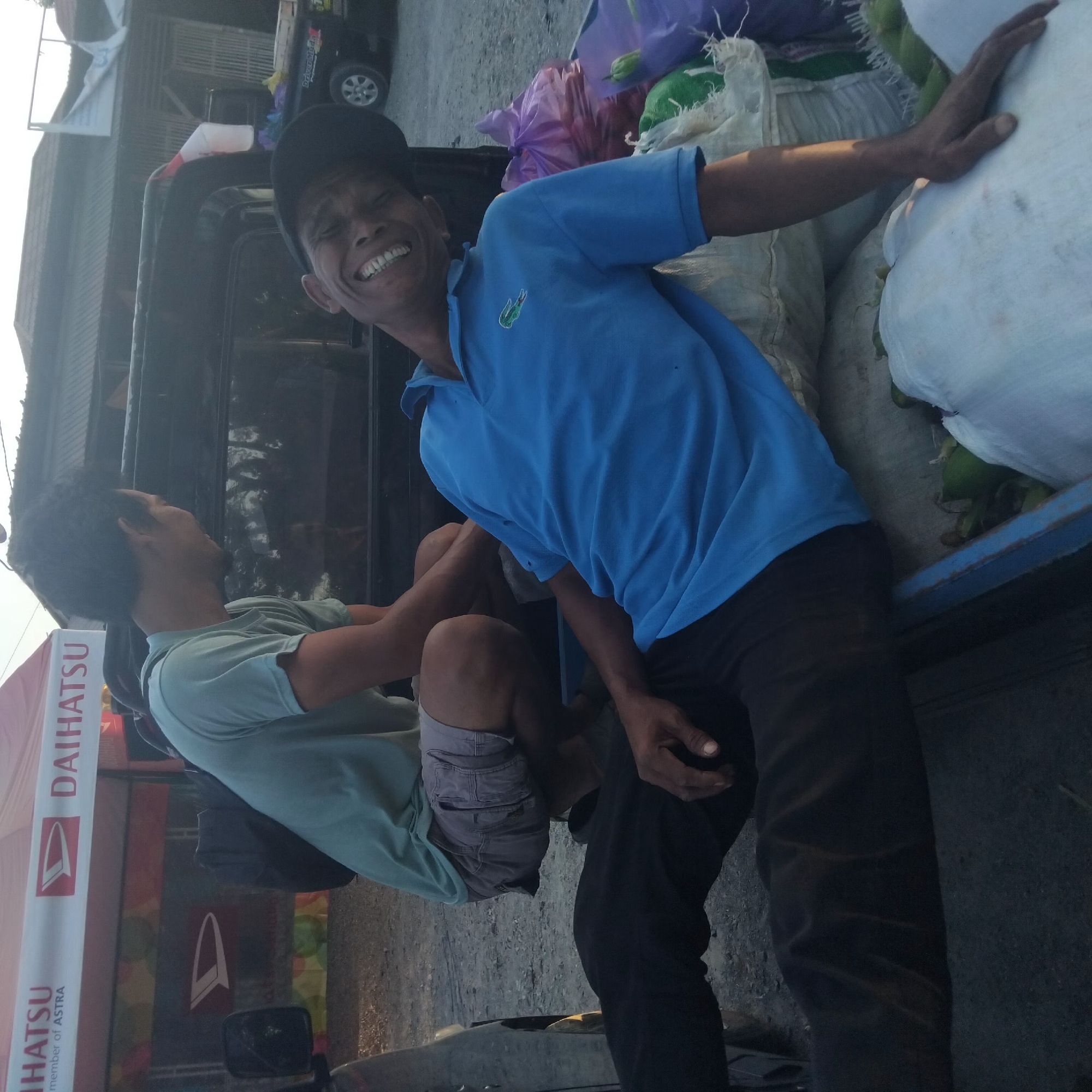 Two men are in the back of an open pickup truck loaded with produce. The man in the foreground, wearing a blue polo shirt and a black baseball cap, is lying on his back and smiling broadly with his mouth open, showing his teeth. He is wearing dark pants. The other man, wearing a light green t-shirt and gray shorts, is sitting and facing away from the camera. He has a backpack on. In the background, there is a large tent with "DAIHATSU" branding, suggesting a promotional event or dealership. The setting appears to be an outdoor area, possibly a market or a roadside, with gravelly ground and buildings visible in the distance. The lighting suggests it is either early morning or late afternoon. The produce in the truck appears to be a mix of vegetables, possibly chilies and corn, contained in white, green, and purple bags. The overall scene conveys a casual and possibly humorous interaction between the two individuals. The scene is likely located in Pelaihari, Indonesia, given the Daihatsu branding which is prominent in that region.