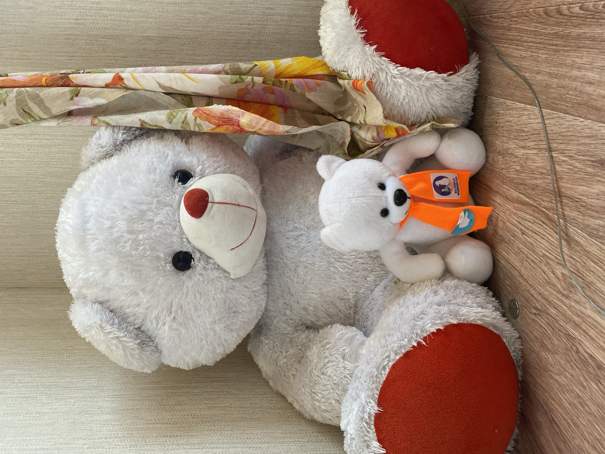 Two teddy bears are sitting on a wooden floor in front of a textured beige wall. The larger bear is white with red paws and a red nose. A small, white teddy bear with an orange scarf is sitting in front of the larger bear. The scarf has a logo with a blue circle and text in Russian. A patterned cloth is draped behind the bears. A coin is on the floor between the bears' feet.