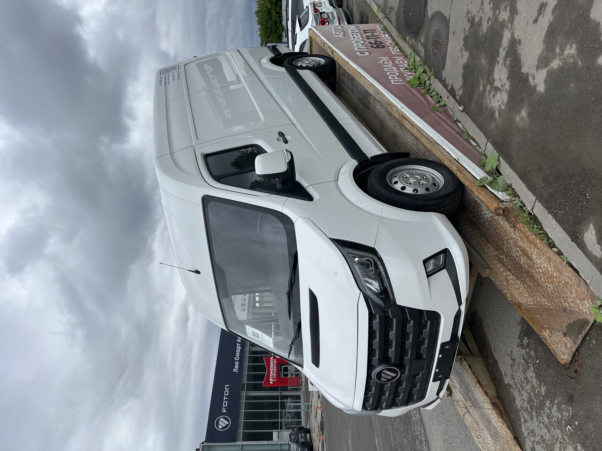 A white Foton cargo van is prominently displayed on rusty metal ramps, creating an upward incline with its front wheels positioned just off the ground and its rear wheels elevated on the ramps. The van appears new, featuring a large black grille with the Foton logo, modern LED-style headlights, and a visible antenna on its roof. Reflections of the dealership building and other elements are visible on the van's side panels.
A red banner covers the side of the display ramps, displaying white Cyrillic text that translates to: "АВТО С ПРОБЕГОМ" (Used Cars), "ПРОДАЕМ" (We sell), a phone number "66-11-11", and "ПОКУПАЕМ ЛЕО СМАРТ АВТО" (We buy, Leo Smart Auto). Weeds are visible growing along the concrete curb adjacent to the ramps.
In the background, a modern Foton dealership building is visible, featuring a glass facade and a prominent "FOTON" logo with "Лео Смарт Авто" (Leo Smart Auto) signage above the main entrance. Inside the showroom, a red sign advertises "АВТОМОБИЛИ С ПРОБЕГОМ" (Used Cars). A dark Foton pickup truck is visible inside the showroom to the left, and several white vehicles are parked in the lot beyond the van. A white sticker on the upper right rear of the van explicitly states "ЛЕО-СМАРТ АВТО", "г. Ижевск, ул. Воткинское шоссе, 182А" (Izhevsk, Votkinskoe shosse, 182A).
The scene is under an overcast, grey sky with numerous clouds. The ground is asphalt pavement, displaying patches of dampness. No people are visible within the frame. The precise location is confirmed as a Foton dealership operated by Leo Smart Auto at Votkinskoe shosse, 182A, Izhevsk, Udmurt Republic, Russia.