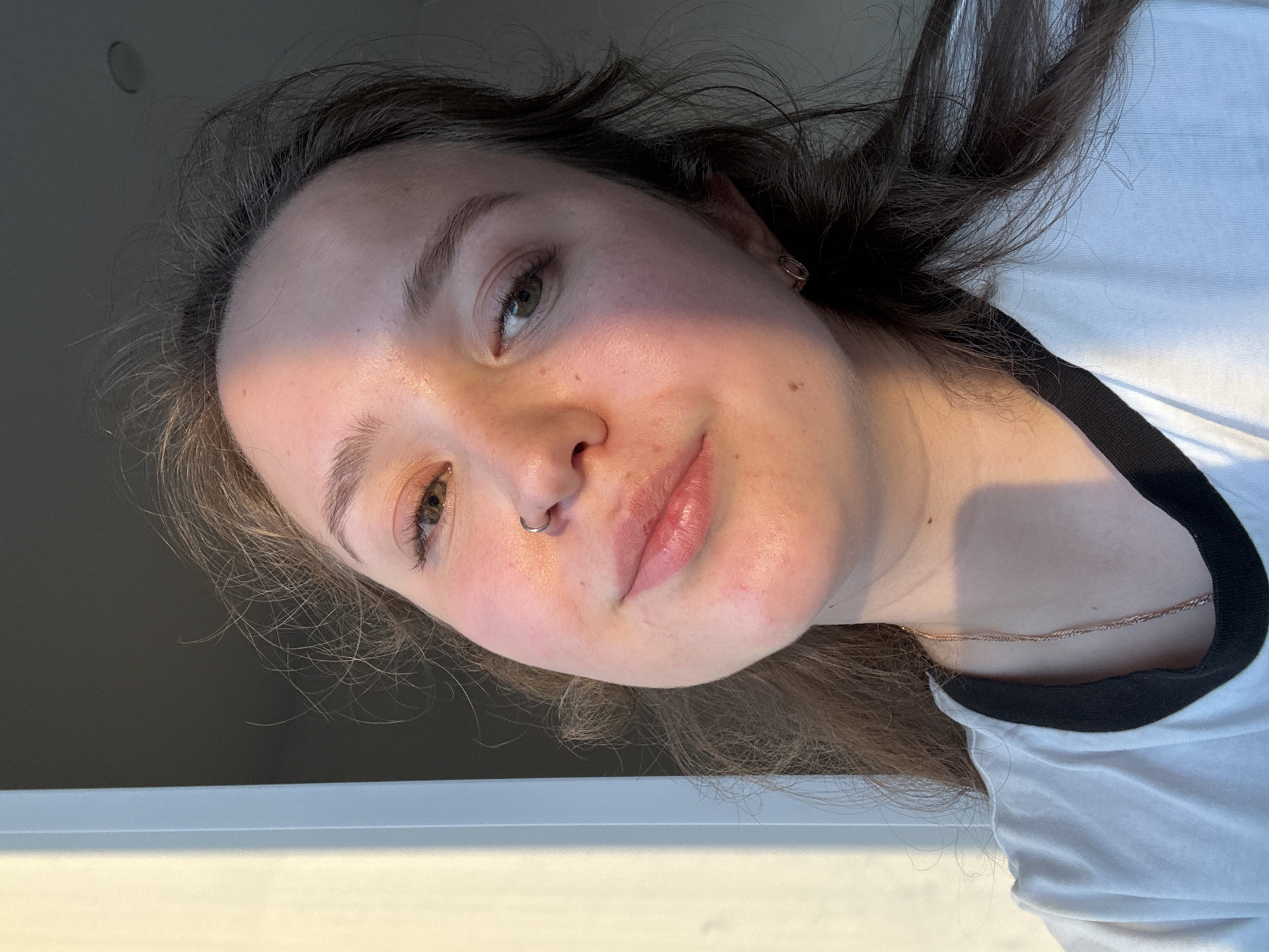 A young woman is captured in a close-up selfie. Her face is slightly angled, revealing details such as a nose ring and light makeup. She has light skin, brown hair, and brown eyes. She wears a white t-shirt with a black collar and a delicate necklace. The background is blurred. The location in Izhevsk, Russia, cannot be determined from the provided image.