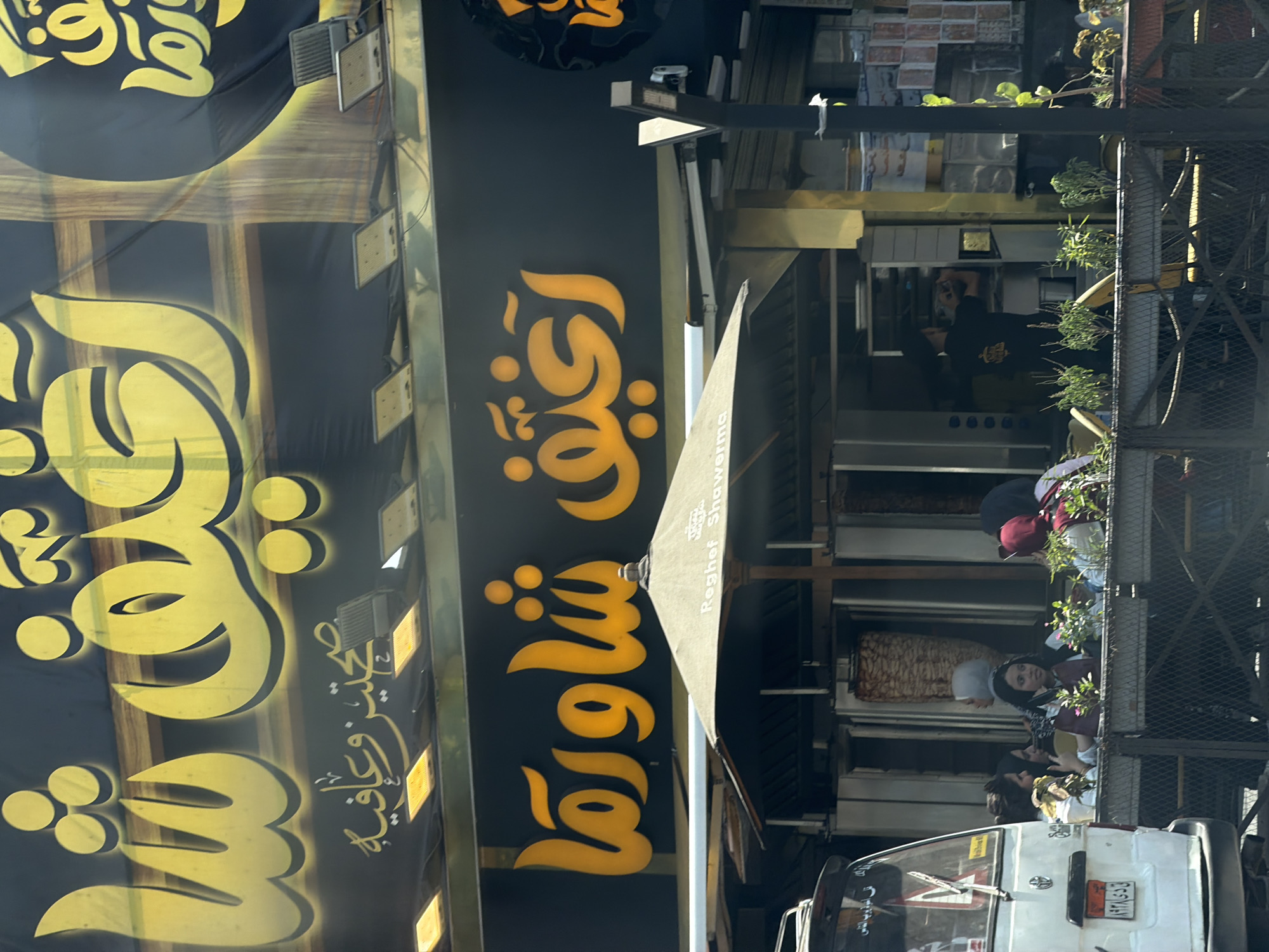 The image captures a storefront for "Raghet Shawarma," a restaurant specializing in shawarma, located in Cairo, Egypt. The prominent signage features large, bright yellow Arabic lettering against a black background, advertising the establishment's name and offerings. Below the main sign, a smaller banner displays more Arabic text, likely a slogan or additional information.
In front of the restaurant, a large outdoor seating area is visible, shaded by a beige umbrella with the restaurant's name printed on it. Several patrons, predominantly women, are seated at tables, engaged in conversation and eating. One person, possibly an employee, is seen behind the counter within the restaurant, attending to customers or preparing food. The shawarma rotisseries are visible, with meat rotating on spits, indicating the food being prepared.
To the left of the storefront, a white van is parked, with its license plate clearly visible. The overall scene suggests a busy, daytime operation of a popular local eatery in a bustling urban environment. The architecture and the style of the signage are consistent with typical establishments found in Cairo. The presence of plants in planters adds a touch of greenery to the outdoor seating area.