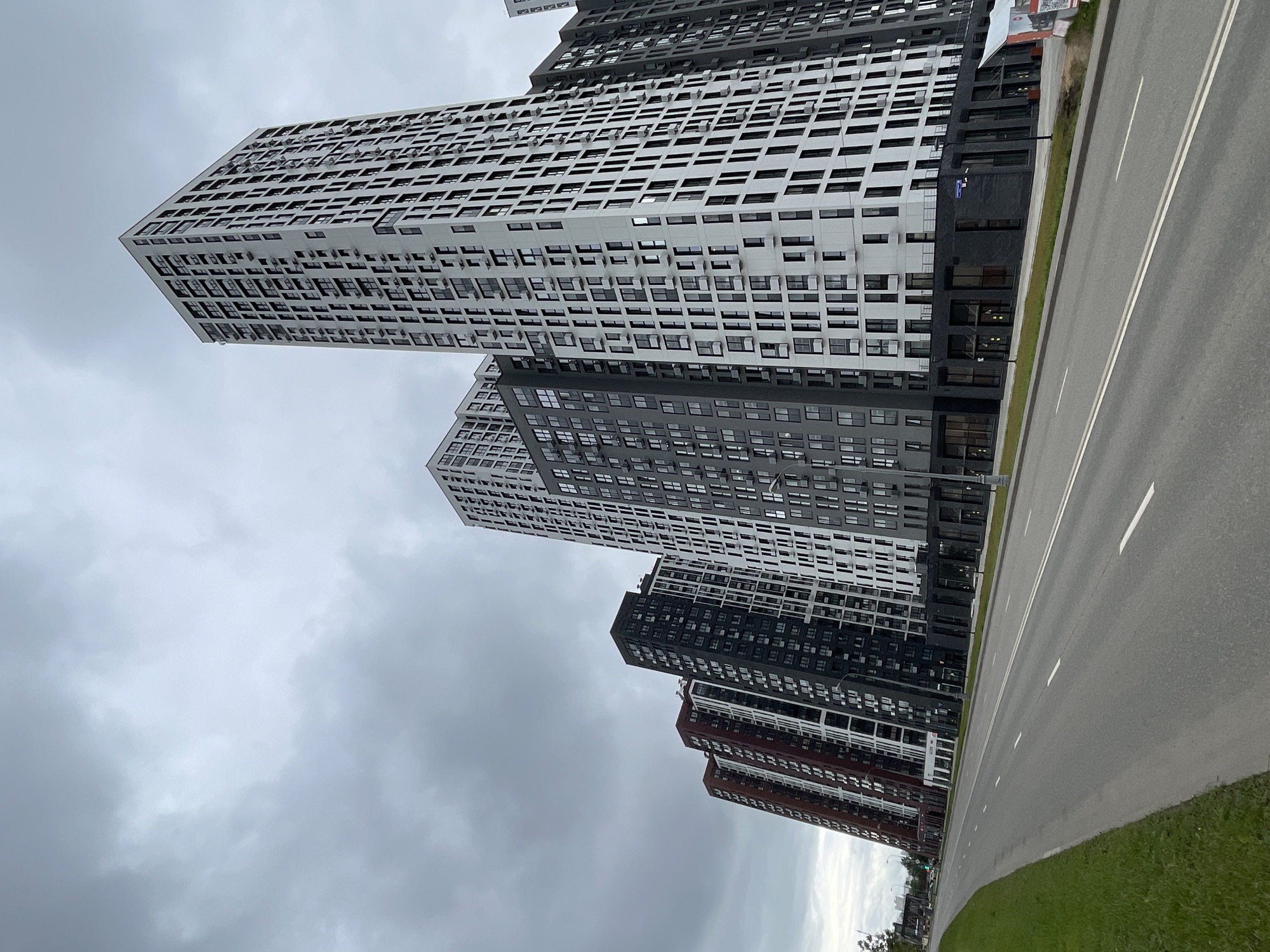 The image shows a row of modern, high-rise apartment buildings in Moscow, Russia. The buildings are tall and slender, with a predominantly white and gray facade, punctuated by numerous windows. The architecture is contemporary, with clean lines and a modular design. In the foreground, a wide asphalt road with white lane markings runs parallel to the buildings. To the left of the road, a strip of green grass and a few trees suggest an urban park or green space. The sky is overcast with heavy, gray clouds, indicating a possibly cool or rainy day. There are no people or vehicles visible in the immediate scene, giving the impression of a quiet, modern urban environment. The buildings appear to be part of a newly developed residential complex.