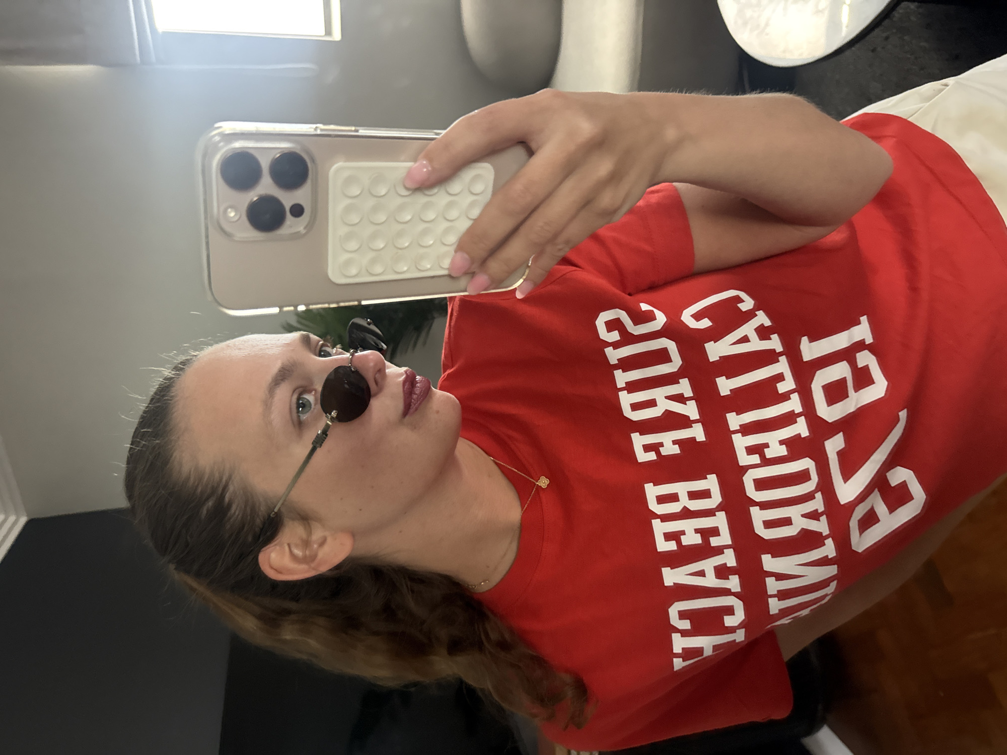 A young woman is taking a selfie indoors. She is wearing a red t-shirt with white lettering that reads "California Surf Beach Like Beyond". She is also wearing round sunglasses with thin frames and a delicate gold necklace with a small pendant. She is holding a smartphone with a clear case that has a white pop-socket style accessory on the back. The background includes a light-colored wall, a dark wall, and a glimpse of a round white table or light fixture. There is no evidence to suggest this image was taken in Cape Town, South Africa.