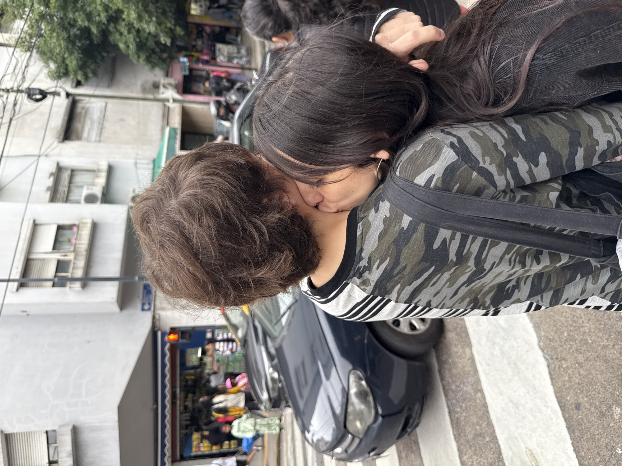 Two individuals are kissing in an urban setting. One person is wearing a camouflage patterned shirt with black stripes on the sleeves and has long dark hair. The other person has short, curly brown hair. They are standing on a crosswalk next to a dark blue car. Behind them is a building with a storefront displaying various items. The location is likely Buenos Aires, Argentina, based on the architectural style and street layout.