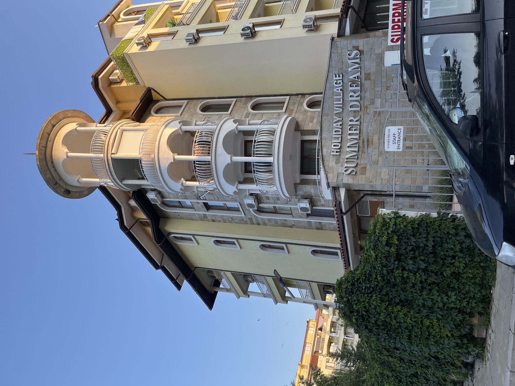 A wide-angle, eye-level, outdoor shot captured in Nesebar, Bulgaria, features a light yellow, multi-story building with a unique curved white balcony structure and a stone facade at its base. The building's architecture is characterized by arched windows, balconies with white railings, and several air conditioning units mounted on the exterior walls. Above the entrance, a stone sign reads "HOLIDAY VILLAGE SUMMER DREAMS." To the left of the entrance, a metal gate is visible with a sign that says "SUMMER DREAMS STAFF ENTRANCE." A lush green ivy-covered wall stands prominently in front of the building, extending towards the left of the frame. In the foreground, the hood and windshield of a black car are partially visible, suggesting it is parked on the street. Another yellow building with balconies is visible behind the ivy-covered wall. The sky is a clear, light blue. The scene appears to be set during a sunny day with the sun casting a warm light on the building.