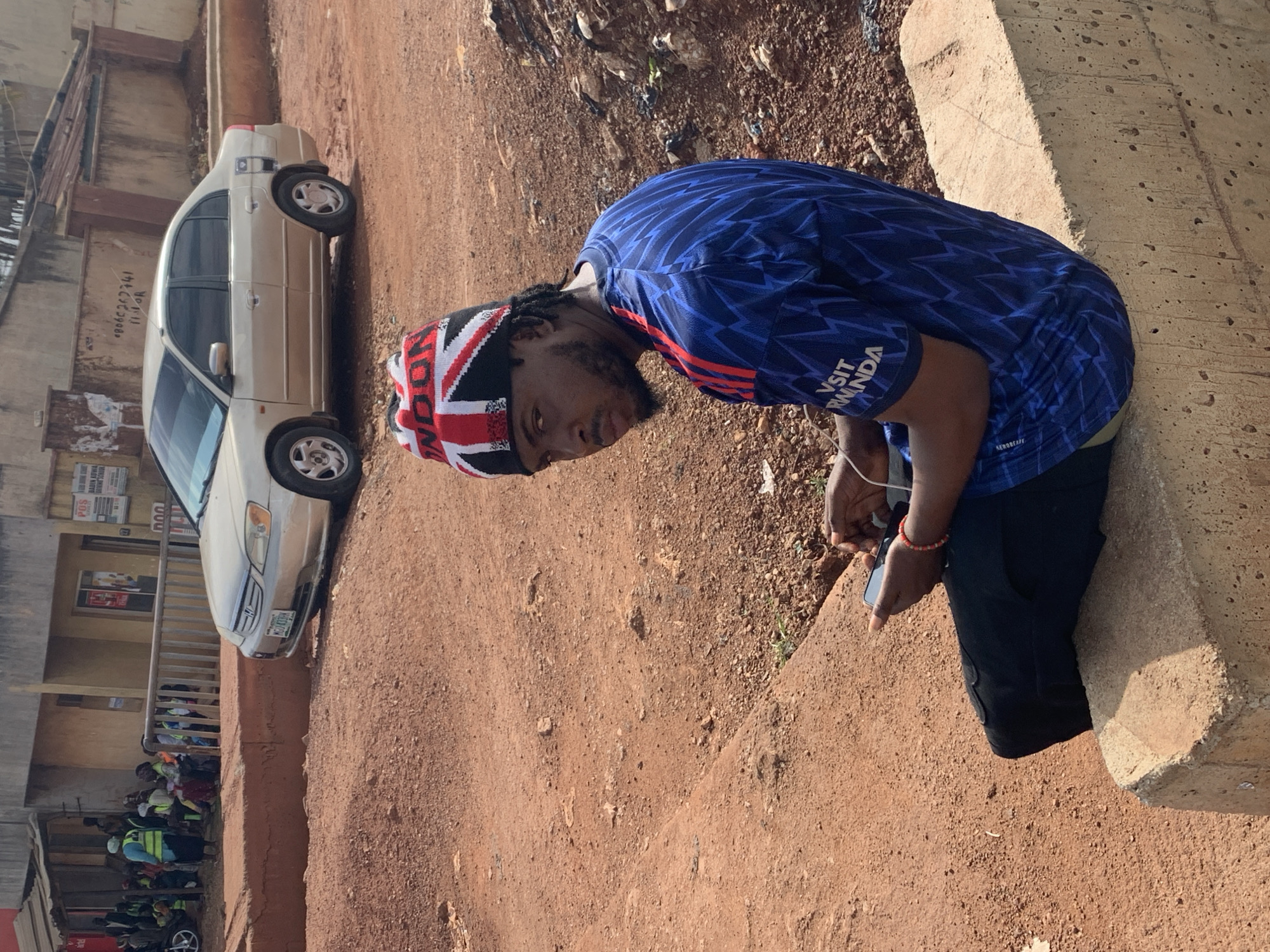 The image captures a street scene in Gura Top, Nigeria. A young man with dark skin, wearing a blue jersey with a geometric pattern and the words "VISIT RWANDA" printed on it, and a beanie featuring the Union Jack flag, sits on a concrete barrier. He is holding a black smartphone in his hands, with white earbuds extending from his ears. His gaze is directed towards the camera. Behind him, a light-colored sedan is parked on a dirt road. In the background, a group of people, some wearing reflective vests, are gathered near a building with a railing. The environment appears to be an outdoor, urban setting characterized by dirt ground and a visible building structure.