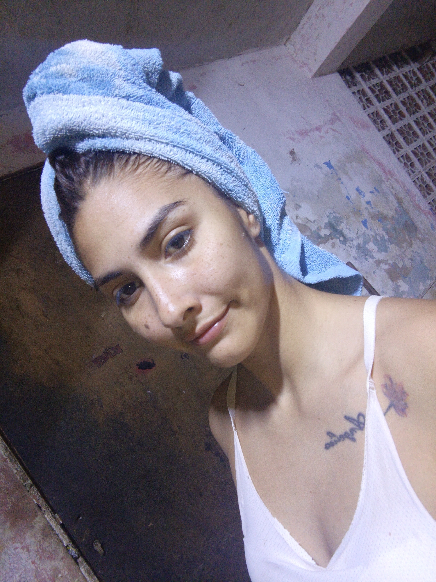 A medium shot captures a young woman indoors. She has wet hair wrapped in a light blue towel. She is wearing a white top with thin straps. Her skin has freckles. She has dark eyebrows and eyes. A tattoo of text is visible on her chest, and another tattoo of a flower is on her shoulder. The background shows a worn wall with peeling paint and what appears to be a patterned vent or window. The scene may be located in Juan Griego, Venezuela based on the image description. A dark door is visible to the left.