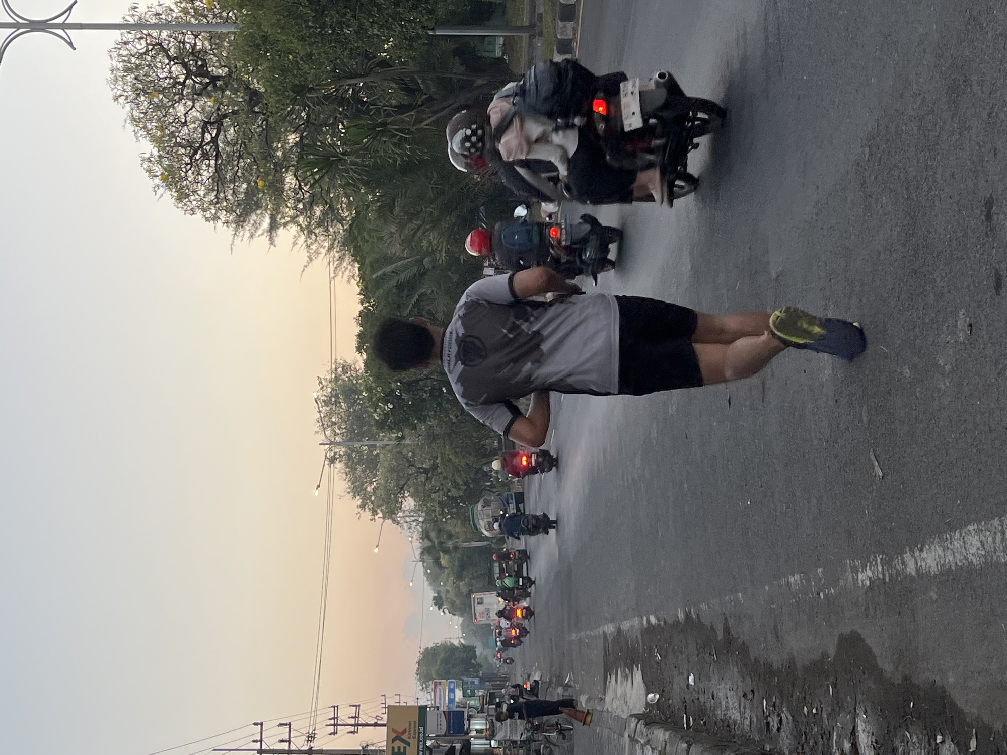 This is a vertical, medium-shot, eye-level, outdoor photograph taken on a paved road in Boro, Indonesia. The scene captures a person running on the right side of the road, facing away from the camera and towards the distance. The runner is wearing a grey and black t-shirt with a graphic design and black shorts. They are also wearing blue and yellow running shoes. Behind the runner, there are several motorbikes traveling in the same direction. On one of the motorbikes, a person wearing a red helmet and a backpack is visible. On another motorbike, two people are riding, one wearing a light pink top and a patterned helmet, and the other wearing a black jacket and a red helmet. The road is lined with trees and vegetation on the right side, and buildings and utility poles on the left. The sky is a soft gradient of pale blue and orange, indicating either sunrise or sunset. The overall impression is one of a bustling street scene with urban elements and natural scenery, bathed in the gentle light of dawn or dusk.