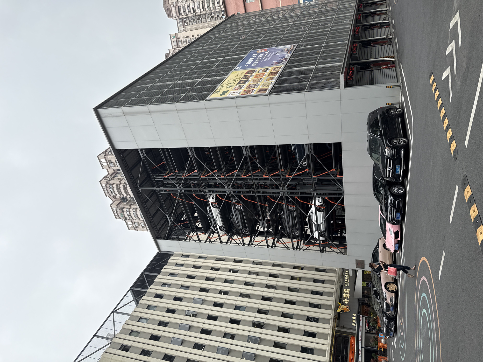 Here's a detailed description of the image:
The image captures an outdoor scene in what appears to be an urban area, likely in Liwan District, China based on the visible storefront signage. The composition is dominated by an automated parking structure, a multi-level steel framework enclosed by a building facade with what looks like a white grid-like facade above and a wire mesh roof on top of that with a large banner of food on it. Multiple cars are parked within this structure.
Adjacent to the parking structure is a street lined with parked cars, including a distinctive pink Lamborghini, and other dark-colored vehicles. A woman is walking along the street next to the parked cars, carrying a red tote bag.
The environment includes other buildings in the background, with light-colored facades and multiple windows. Storefronts are visible with Chinese lettering. The street has colorful, semi-circle markings on the ground. There are also traffic calming devices (speed bumps) along the side of the street. The sky is overcast, suggesting a day with soft, diffused lighting.