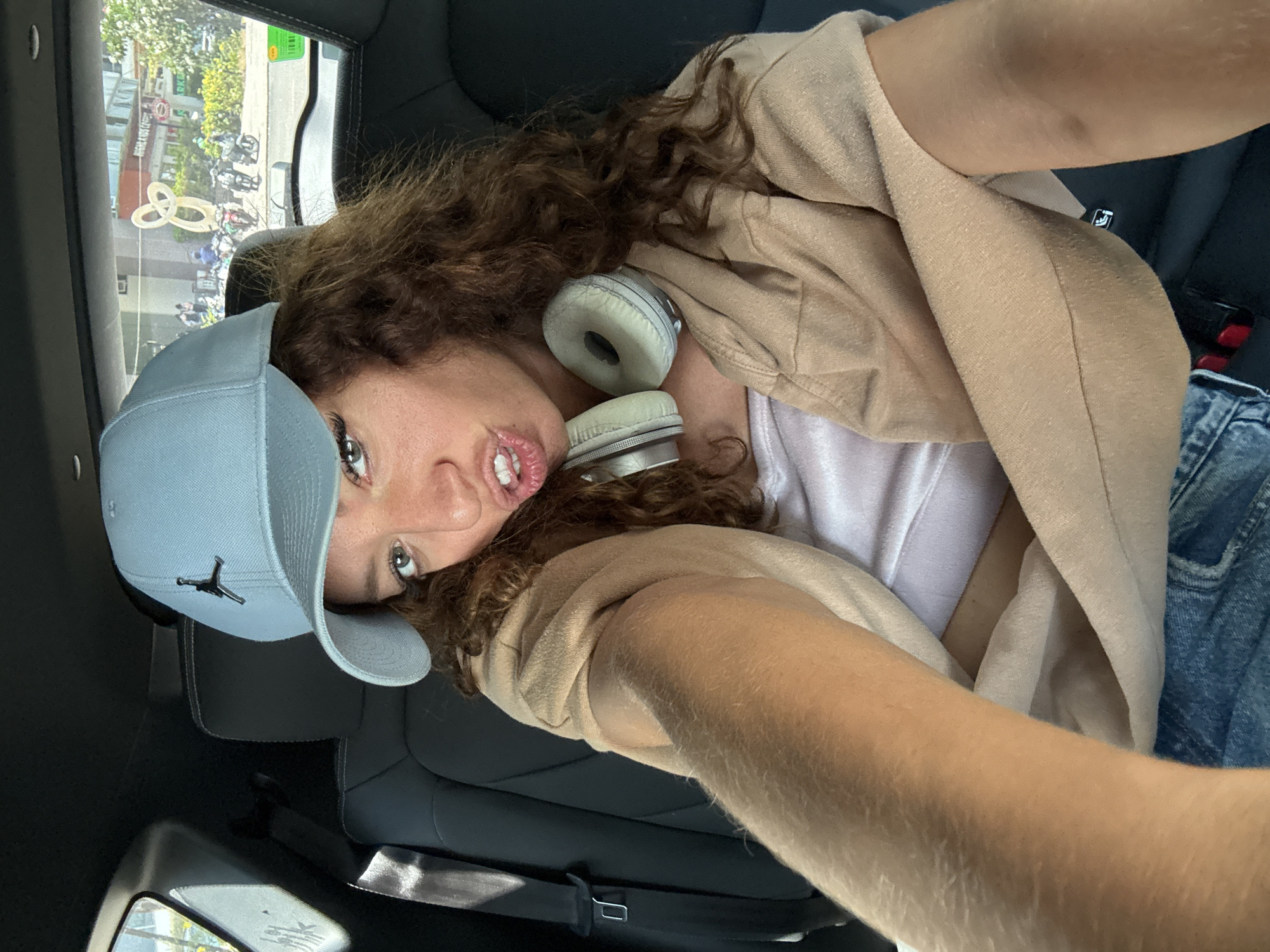 A person is in the back seat of a car. The person is wearing a light blue baseball cap, a light-colored t-shirt, a tan jacket, and light blue denim jeans. White headphones are around the person's neck. The car appears to be stopped, as a street scene is visible through the car window. There are people walking and riding scooters on the street. Buildings and trees are also visible. The location is not visibly verifiable as Đà Nẵng, Vietnam, but it could be any urban area with similar infrastructure.