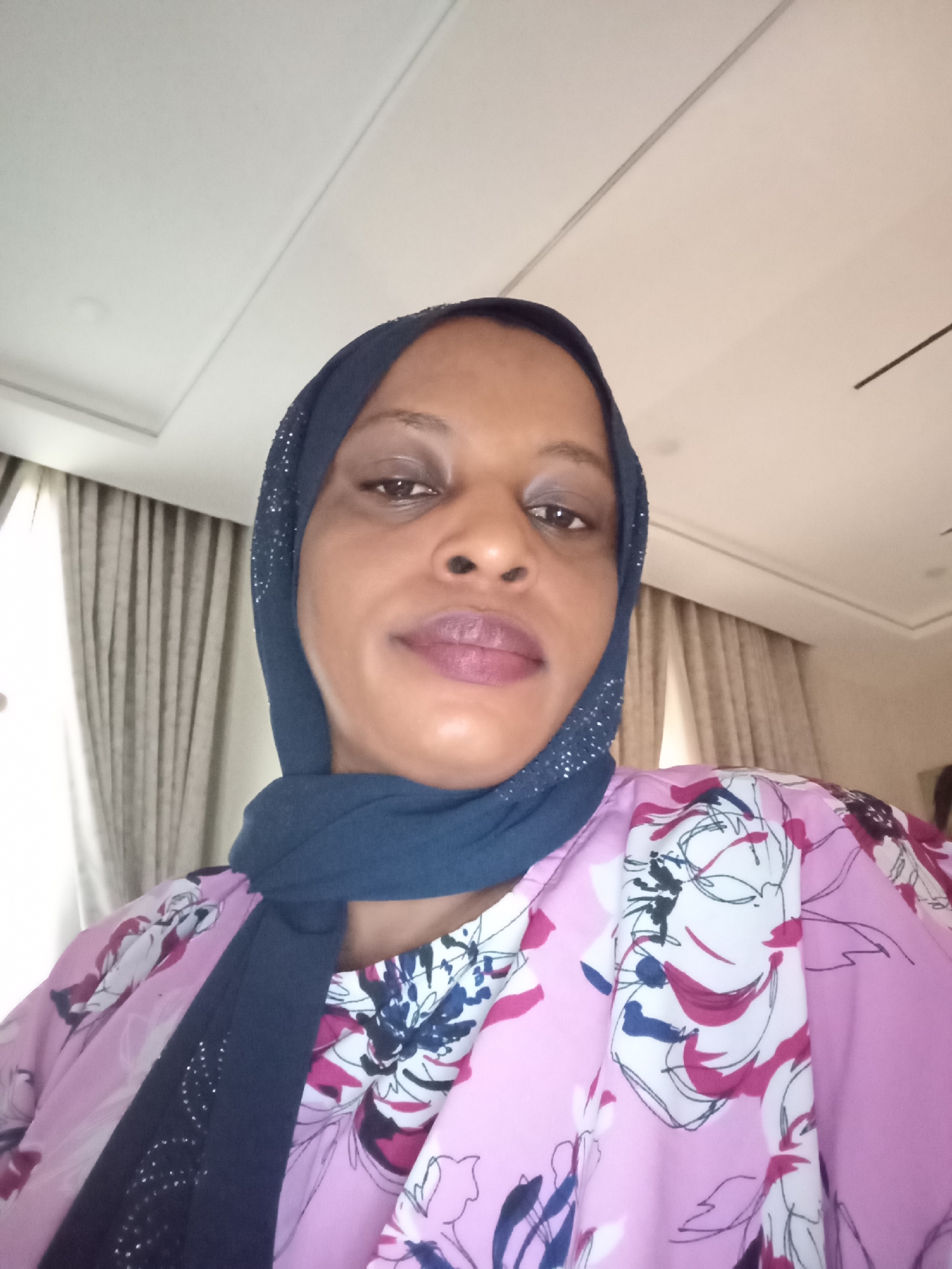 A medium, slightly angled, close-up shot of a woman wearing a dark blue hijab that is lightly decorated with rhinestones around its edge and a pink floral blouse. The setting appears to be an interior space, possibly a room. The background shows neutral-toned curtains, a ceiling with inset rectangular panels, and recessed lighting, suggesting a modern interior. The location is identified as Unguwar Rimi, Nigeria.