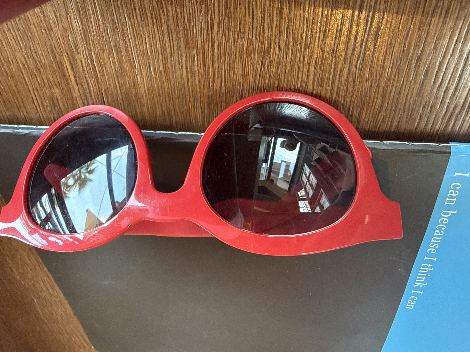 A pair of novelty red heart-shaped sunglasses is placed on top of a dark book that rests on a wooden surface. The sunglasses are positioned diagonally with the top lens reflecting a bright, outdoor scene with sunlight and what appears to be a palm tree. The book has a blue edge along the bottom, with white text that reads "I can because I think I can". The wooden surface shows a distinct wood grain pattern and appears to be a table. There are no people or animals visible in the image. The lighting suggests it is daytime, with sunlight casting reflections on the sunglasses. The scene is static and there are no discernable actions or interactions. The location is not specified within the image.