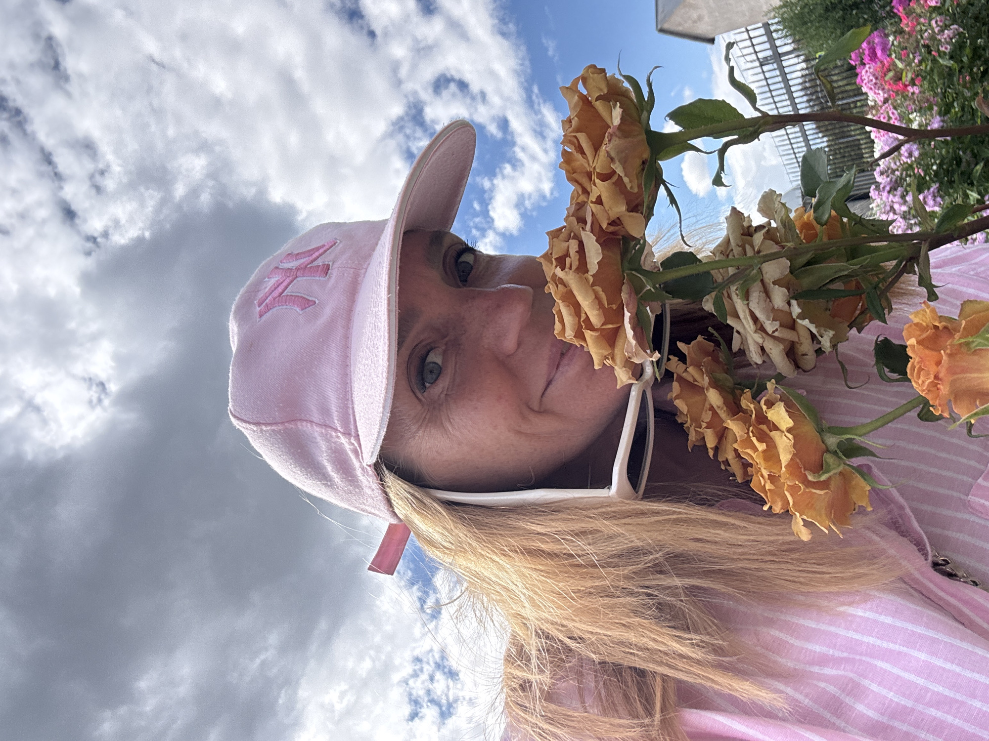 In Saint Petersburg, Russia, a woman wearing a pink baseball cap and a pink striped shirt is captured in a close-up, low-angle selfie. She is smelling a bouquet of orange roses. White-rimmed sunglasses are perched on her shirt collar. Behind her, a sky with dramatic clouds and a hint of blue is visible, along with a garden featuring various flowers and a fence.