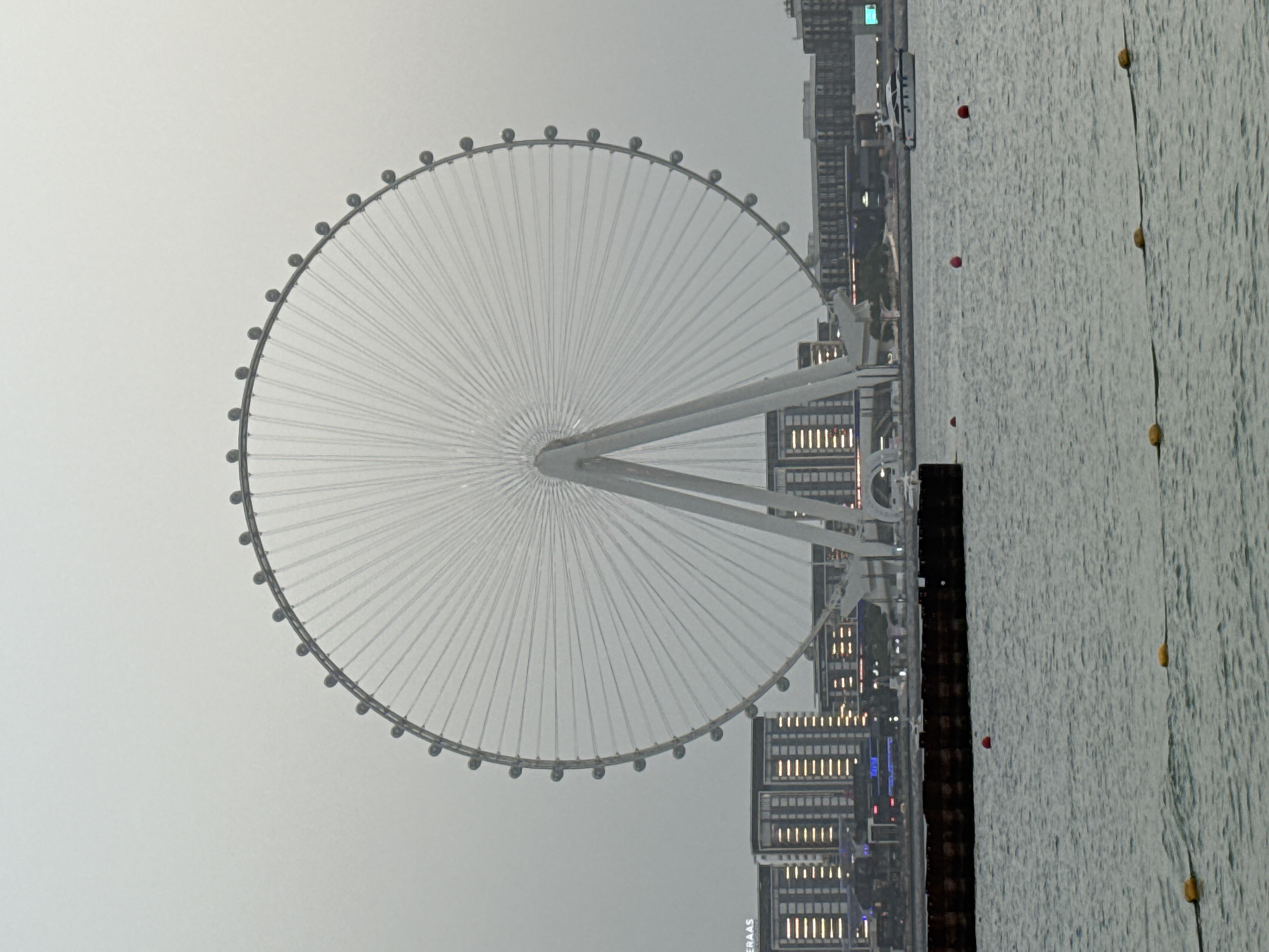 The image shows a Ferris wheel, identified as Ain Dubai, situated on Bluewaters Island in Dubai, United Arab Emirates. The Ferris wheel, a dominant structure, is white and appears to be stationary. It is surrounded by modern buildings and a body of water in the foreground. The sky is overcast, and the overall lighting suggests it is either dawn or dusk. Several red and yellow buoys are visible in the water, and a white yacht is docked near the shoreline in the background. The scene is calm and depicts a city skyline with a prominent landmark.