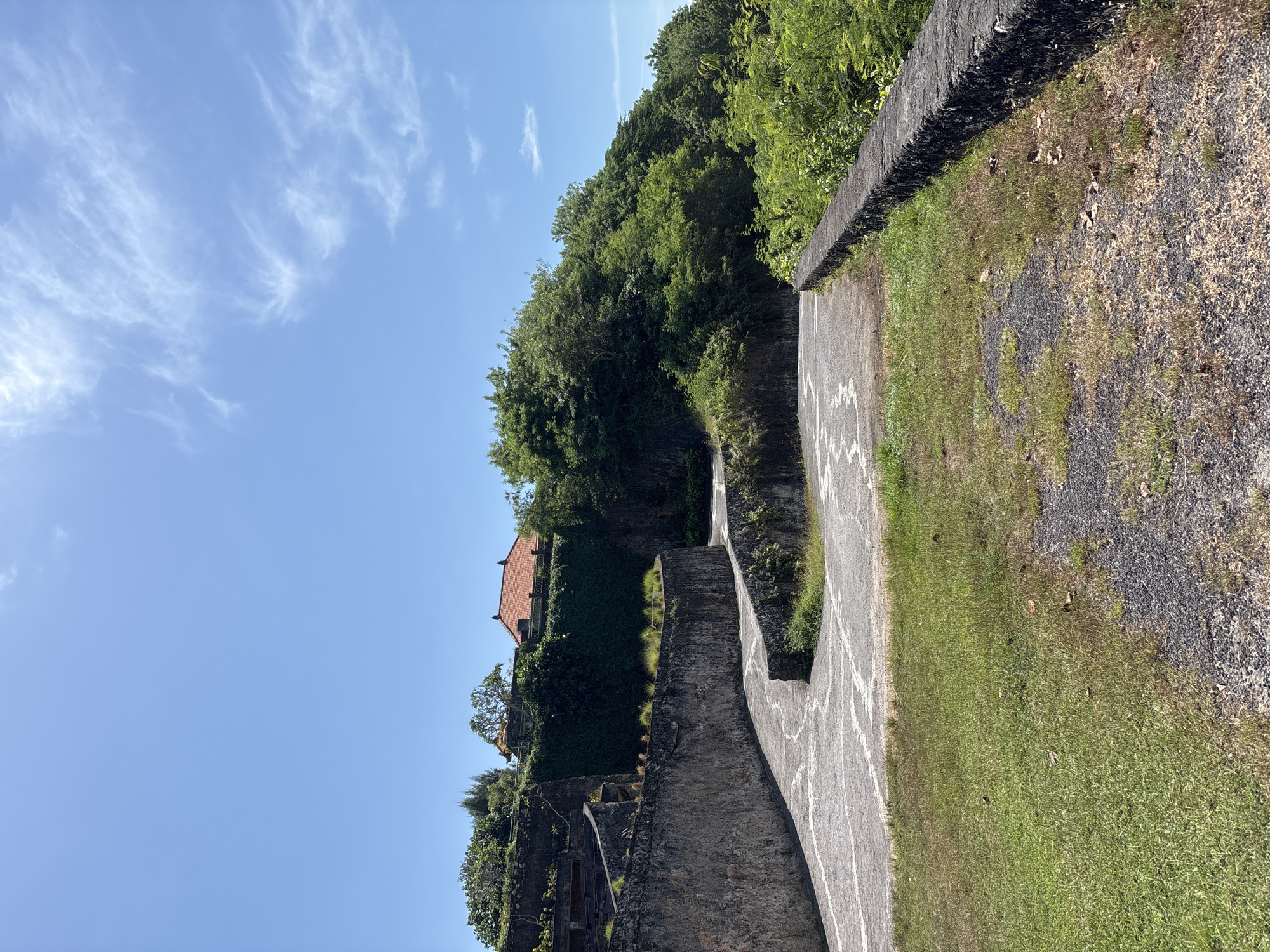 The image, presented in a rotated vertical format, captures a bright outdoor scene featuring a concrete pathway winding alongside a verdant hillside under a clear blue sky.
When viewed horizontally, the pathway occupies the foreground and mid-ground, extending into the distance. It features faint, stylized white markings on its surface. To the left of the path, a stone wall separates it from a patch of ground covered in a mix of green grass, dry earth, and small stones.
On the right side of the pathway, a steep incline is densely covered with lush green trees and tropical foliage. A notable feature nestled within this greenery is a building with a distinctive red-tiled roof, partially visible through the leaves. Further along the hillside, other stone structures or terraced elements can be discerned amidst the vegetation.
The upper portion of the frame is dominated by a clear, bright blue sky adorned with scattered, wispy white clouds, indicative of sunny weather. The strong natural light suggests it is daytime, likely mid-morning or early afternoon.
No people or discernible activities are taking place, and there is no visible text. The overall scene evokes a tranquil, natural environment, consistent with the tropical setting of Kuta Selatan, Indonesia, possibly depicting a path through a park, resort grounds, or a scenic cliffside walk.