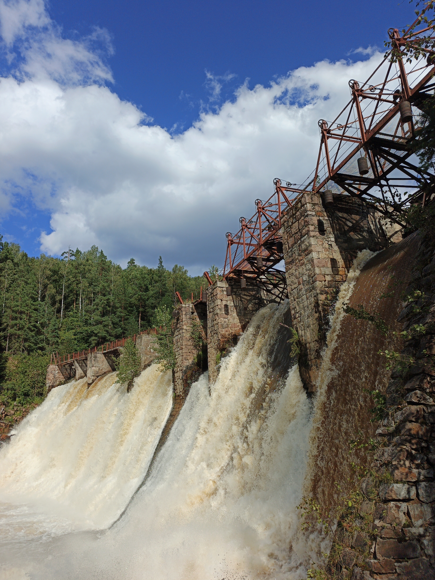 The image depicts the Porogi Hydroelectric Station, a historic structure located in Porogi, Russia. The dam is characterized by its stone construction and a prominent red metal superstructure with intricate mechanisms, likely for controlling water flow. Powerful cascades of water are plunging down the dam's face, creating a dramatic scene. The surrounding environment is lush and green, with a dense forest visible on the hillside behind the dam. The sky is a vibrant blue, punctuated by scattered white clouds. The perspective is from a low angle, looking up towards the dam and the sky. There are no people visible in the image. The overall impression is one of industrial heritage set against a natural, powerful landscape.
