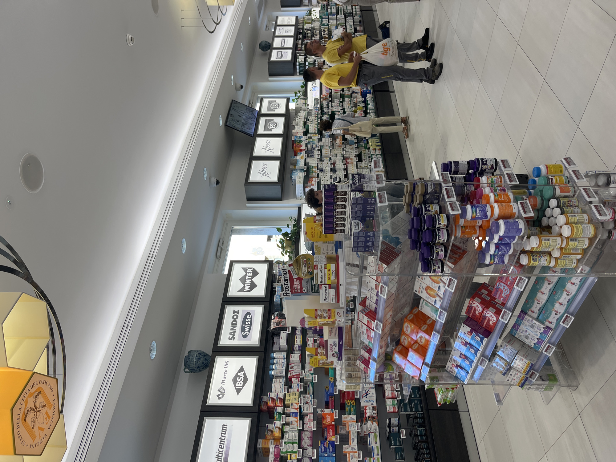 The image captures the interior of a pharmacy located in Città del Vaticano, Vatican City. The pharmacy displays a wide array of pharmaceutical products and supplements on shelves and tiered racks. Prominently visible are illuminated signs above the shelves featuring brand names such as "Multicentrum," "IBSA," "Sandoz," and "Winter." Several customers are browsing the aisles, with two men in yellow t-shirts standing near a checkout counter, one holding a shopping bag. The environment is well-lit, with modern lighting fixtures and a tiled floor. A decorative vase is positioned on a high shelf. The overall scene depicts a typical retail environment within a pharmacy.