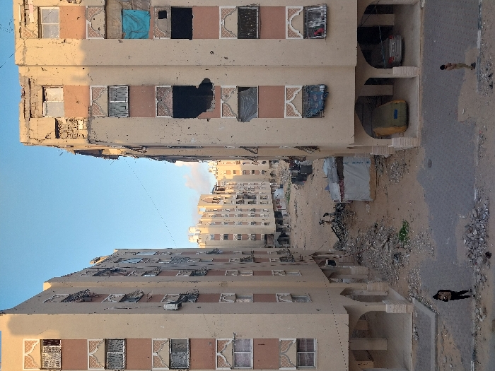 The image depicts a damaged urban street in Khan Yunis, Palestinian Territory, viewed from an elevated perspective. Two multi-story residential buildings, primarily light beige in color, flank a street that extends into the distance, revealing more similar structures. Both foreground buildings exhibit extensive damage, including numerous impact marks consistent with bullet holes across their facades, shattered or missing windowpanes, and significant structural compromise, particularly at their upper levels where rebar is exposed.
On the right building, several windows are covered with blue and black tarp-like materials. The ground level of both buildings features arched openings. Underneath the right building's arches, a yellow object covered by a tarp or fabric, and a silver car, are parked.
The street itself is paved with dark interlocking bricks in the immediate foreground, transitioning to a heavily disturbed dirt and rubble-strewn area further into the scene. Large piles of concrete debris and other construction waste are visible in the middle ground. Amidst this rubble, a small, makeshift structure covered in white tarp material stands.
Four individuals are present. In the paved foreground, one person in dark clothing and a backpack stands on the left, facing right. Another person in light-colored clothing stands on the right, facing left. In the middle ground, near the makeshift structure and rubble, two smaller individuals are observed, appearing to interact with the debris. The sky above is clear and light blue, indicating daytime conditions.